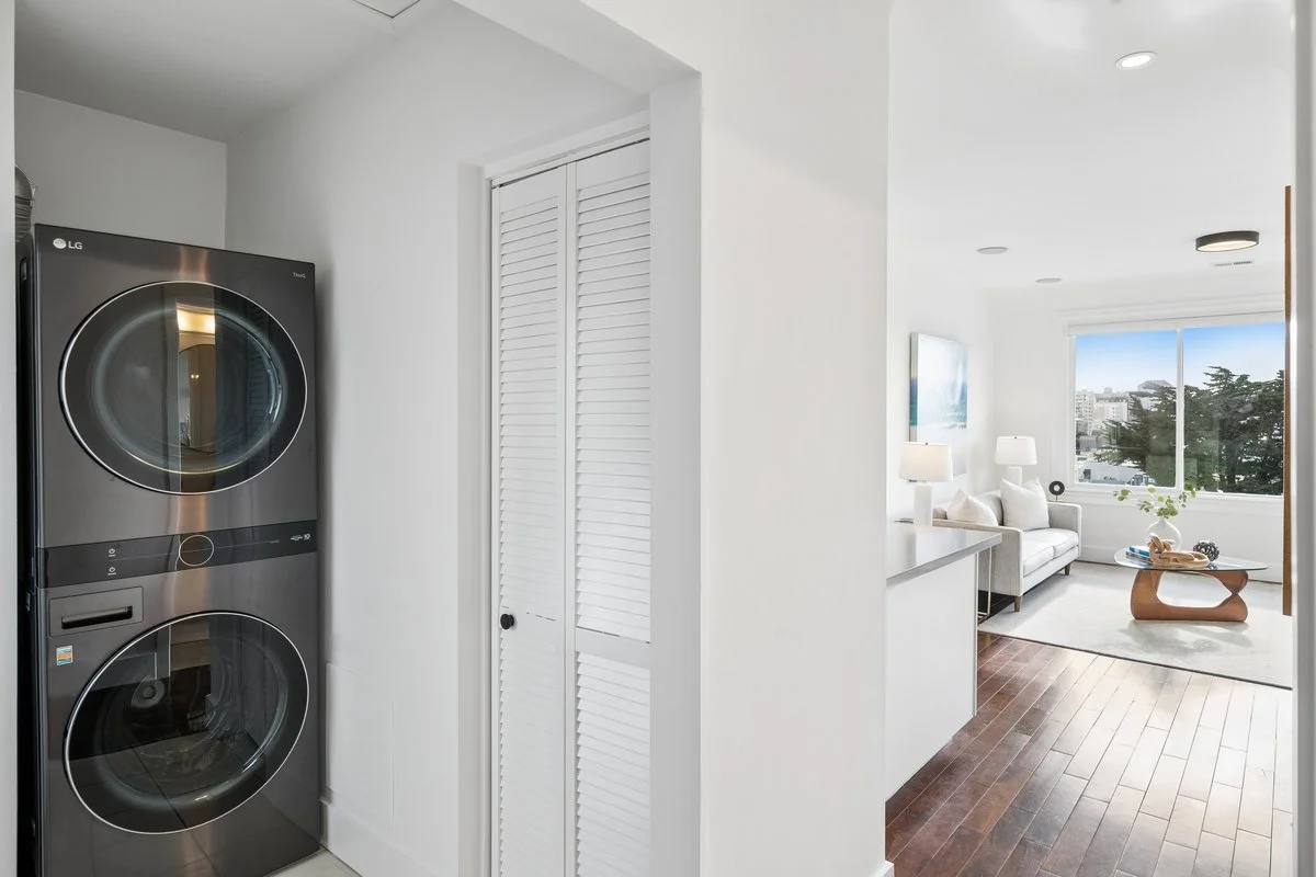 Laundry area with a stacked LG washing machine and dryer, adjacent to a closet with white louvered doors, opening into a bright living room with white walls, wood flooring, a sofa, lamps, a window, and a coffee table.