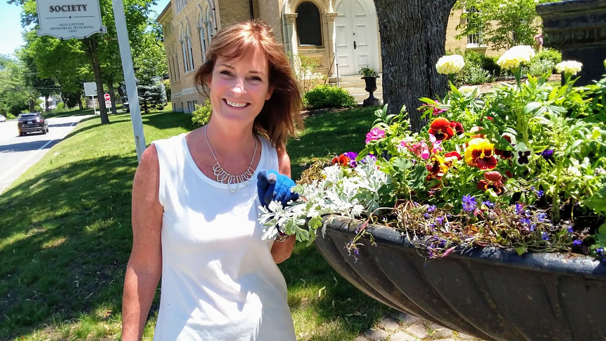 A woman with short, light brown hair wearing a white sleeveless top and a silver necklace, smiling while holding a small plant, standing outdoors next to a large flower planter filled with colorful flowers including white, pink, red, purple, and yell