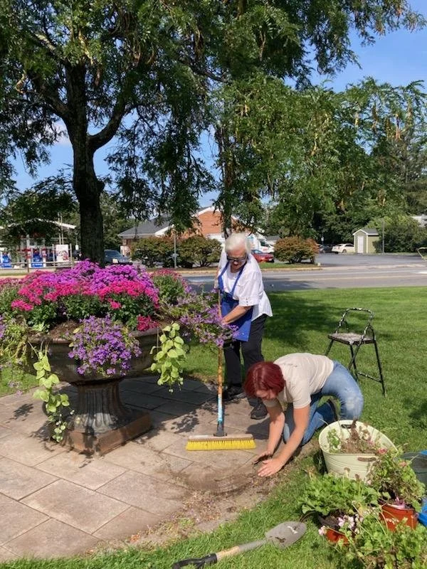 Two women are gardening and planting flowers in a small garden area near a sidewalk with a large flower pot filled with pink and purple flowers. One woman is kneeling and planting, while the other woman stands holding a broom and cleaning the area. There are trees, a street, and houses in the background on a sunny day.