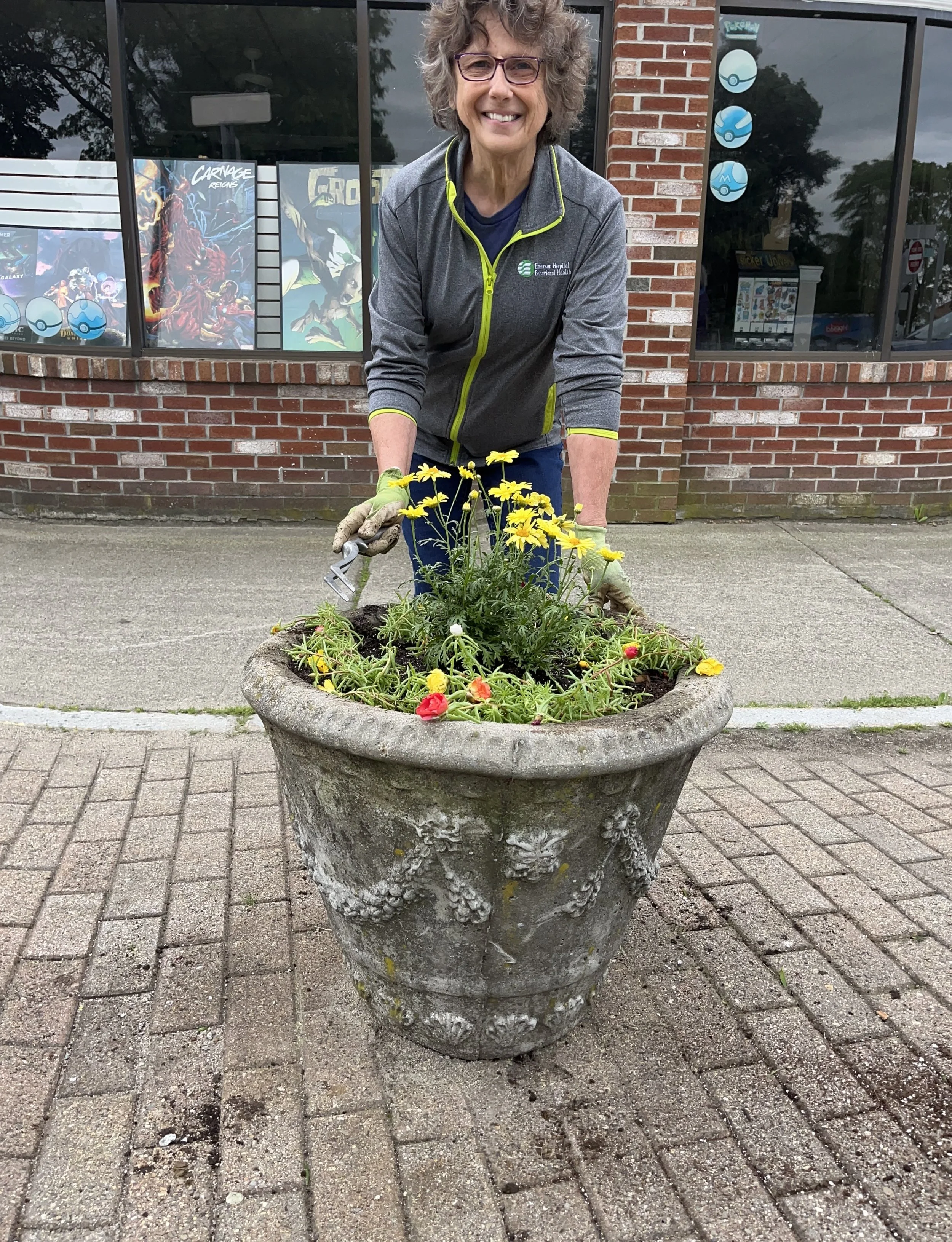 A woman with curly hair, glasses, and a gray jacket kneels in front of a large decorative planters with yellow and orange flowers, smiling at the camera outside a building with brick walls and large windows decorated with Pokemon posters.