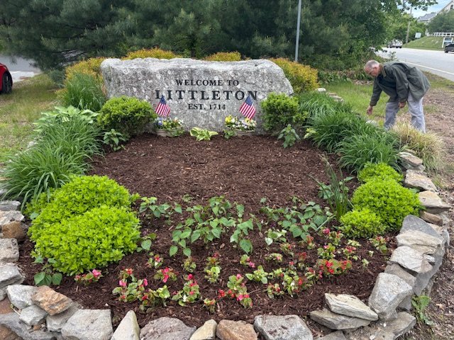 A man planting flowers in a landscaped garden in front of a large rock with a plaque that reads 'Welcome to Littleton Est. 1714,' surrounded by green shrubs and flowering plants.