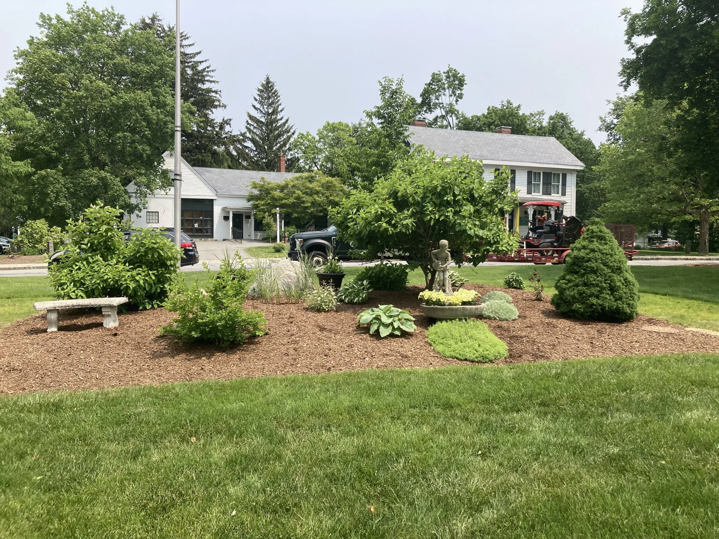 A landscaped garden with various shrubs, a small tree, flower pots, and a garden sculpture in the center. In the background, there are houses and cars parked along the street, with trees and a clear sky.