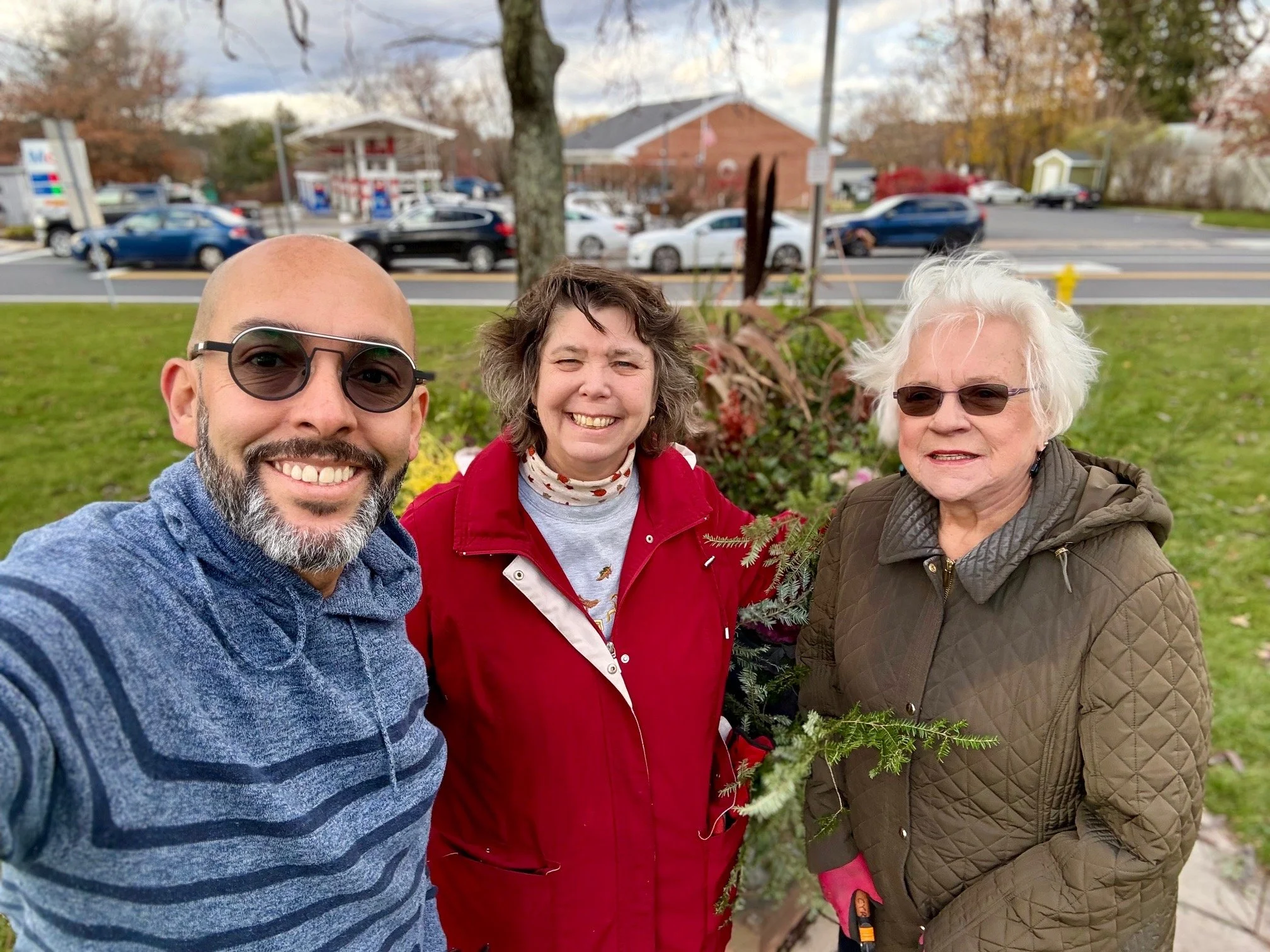 Three people stand outdoors in a park, smiling at the camera. The man on the left is wearing sunglasses and a blue striped sweater. The woman in the middle is wearing a red jacket and a white scarf with red spots. The woman on the right is wearing gl