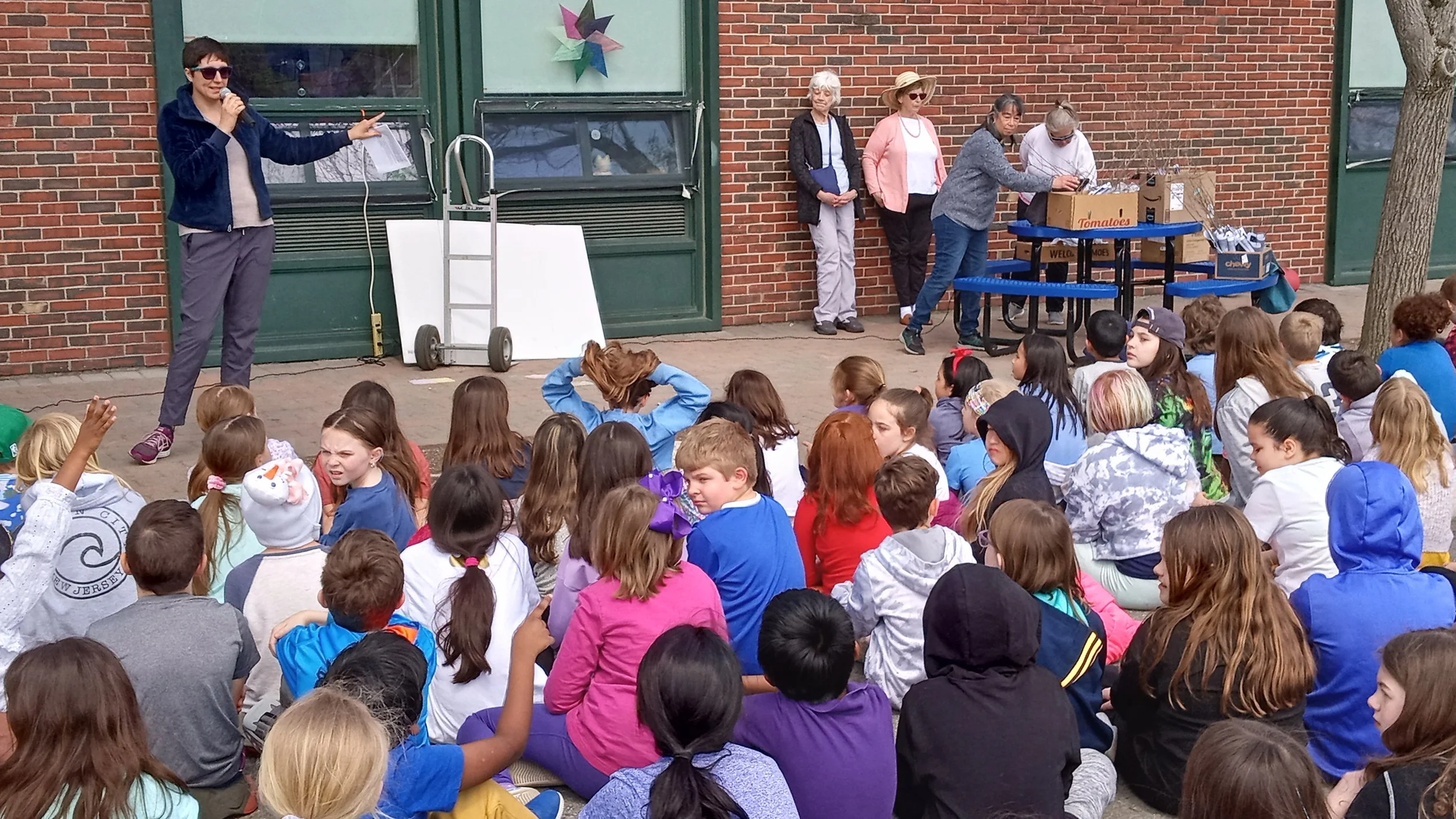 A large group of children sitting on the ground outdoors, listening to a man in sunglasses who is speaking into a microphone and gesturing with his hand. Several adults stand nearby against a brick wall, some behind a table with boxes and some near a tree.