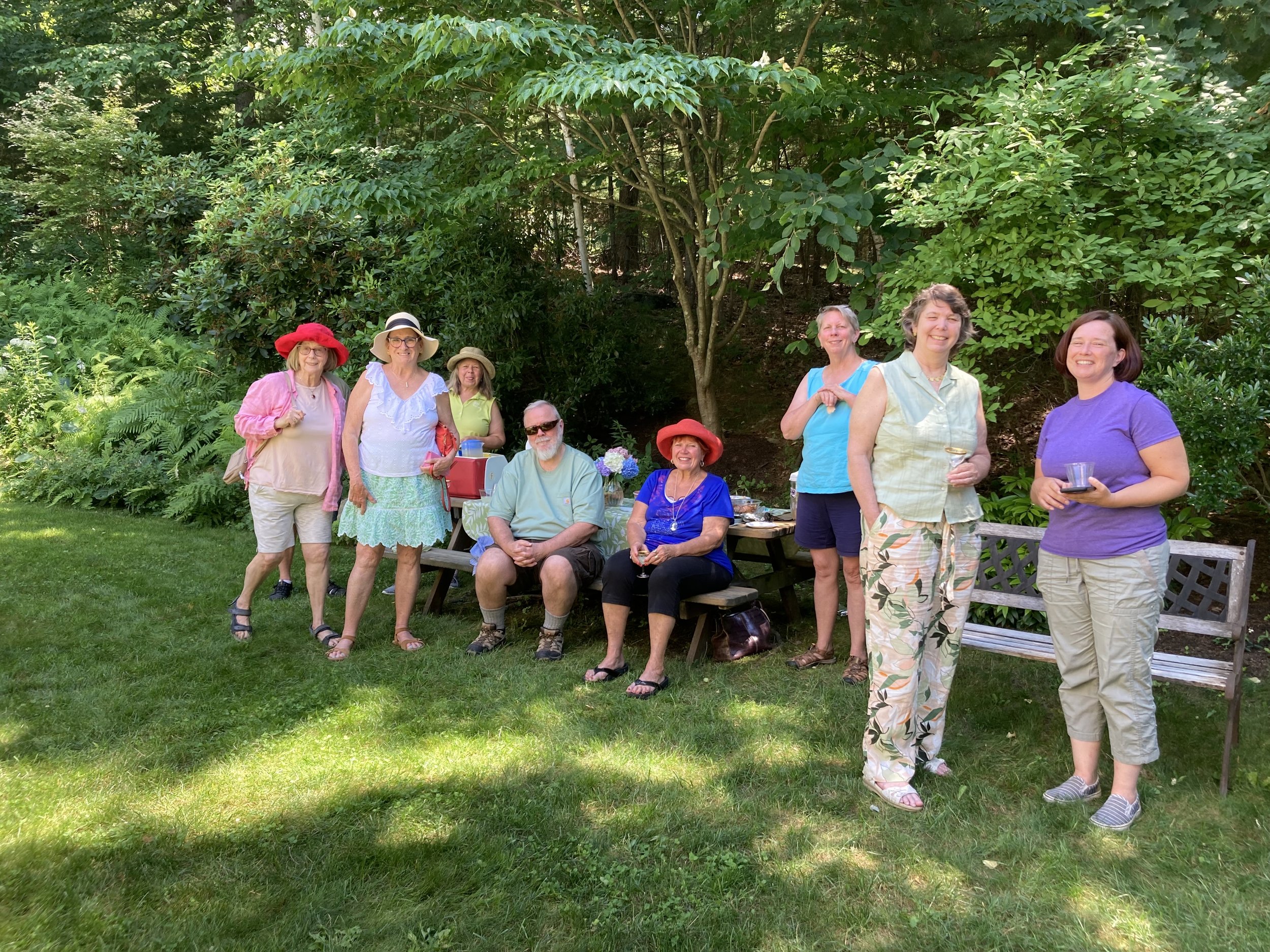 A group of nine people enjoying a gathering outdoors in a green garden, some sitting on a bench and others standing, with trees and bushes in the background.