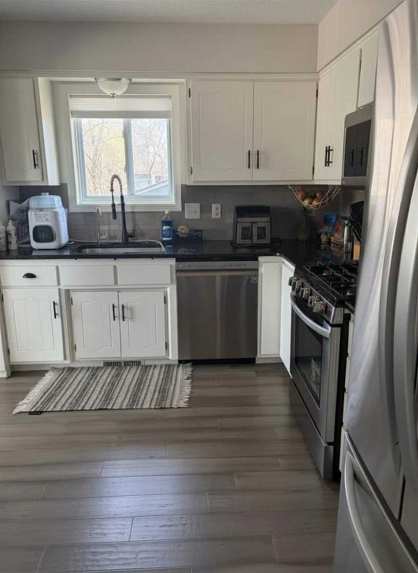 Kitchen with white cabinets, black countertops, stainless steel dishwasher and oven, window above sink, blue soap dispenser, small kitchen appliances, and a striped rug on wood-look flooring.