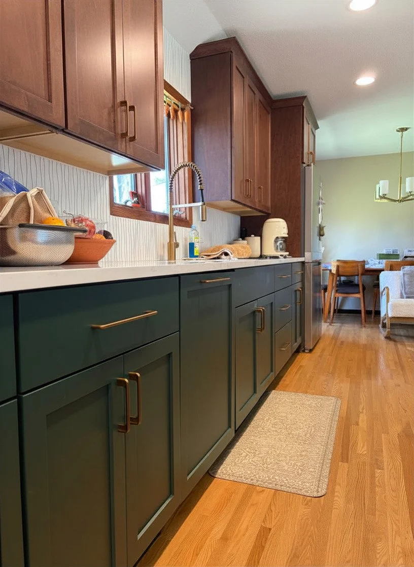 A kitchen with dark green lower cabinets and brown upper cabinets, a white countertop, and hardwood flooring. There is a stainless steel sink with a high-arched faucet and a small window above it. Various kitchen items are on the counter, and there's a dining area with chairs and a table in the background.