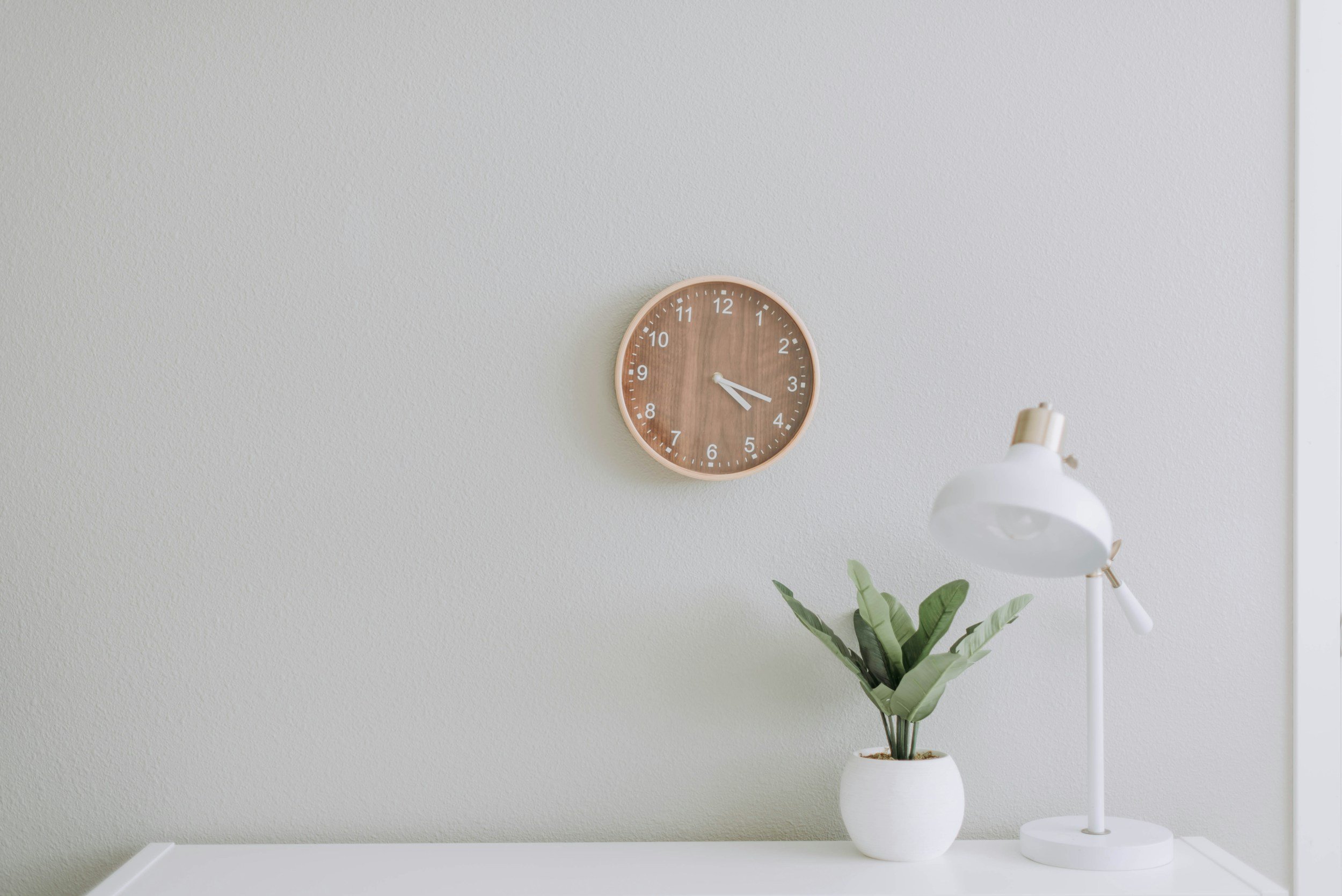 a minimalistic appearance, white dresser top with a flower plant in a round ceraminc vase and a white small lamp by the plant. A minimalistic brown wood wall clock.