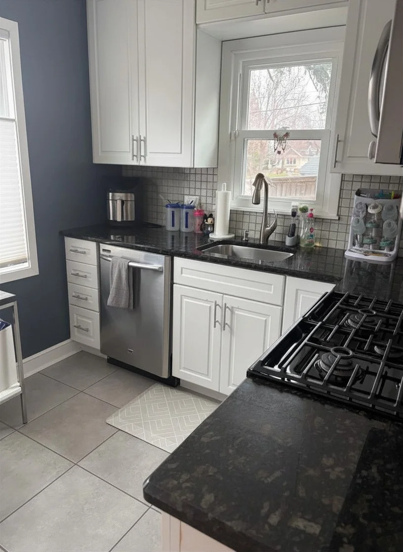 A modern kitchen with white cabinets, a black granite countertop, and a stainless steel dishwasher. There is a window above the sink, which has a faucet and a paper towel roll. Various kitchen items are on the counter, including a coffee maker, containers, and cleaning supplies. The floor has large light gray tiles, and a small patterned rug is in front of the dishwasher.