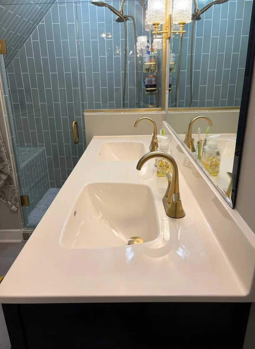 A bathroom vanity with two white sinks, gold faucets, and a large mirror. A shower with blue tiled walls and gold fixtures is visible in the reflection.