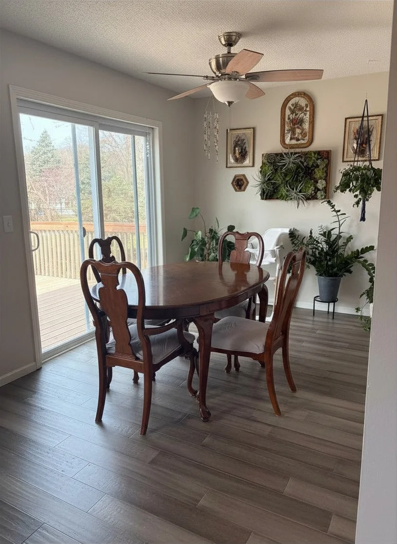 A dining room with a wooden dining table and four chairs, a sliding glass door leading to an outdoor deck, potted plants, wall art, and a ceiling fan.