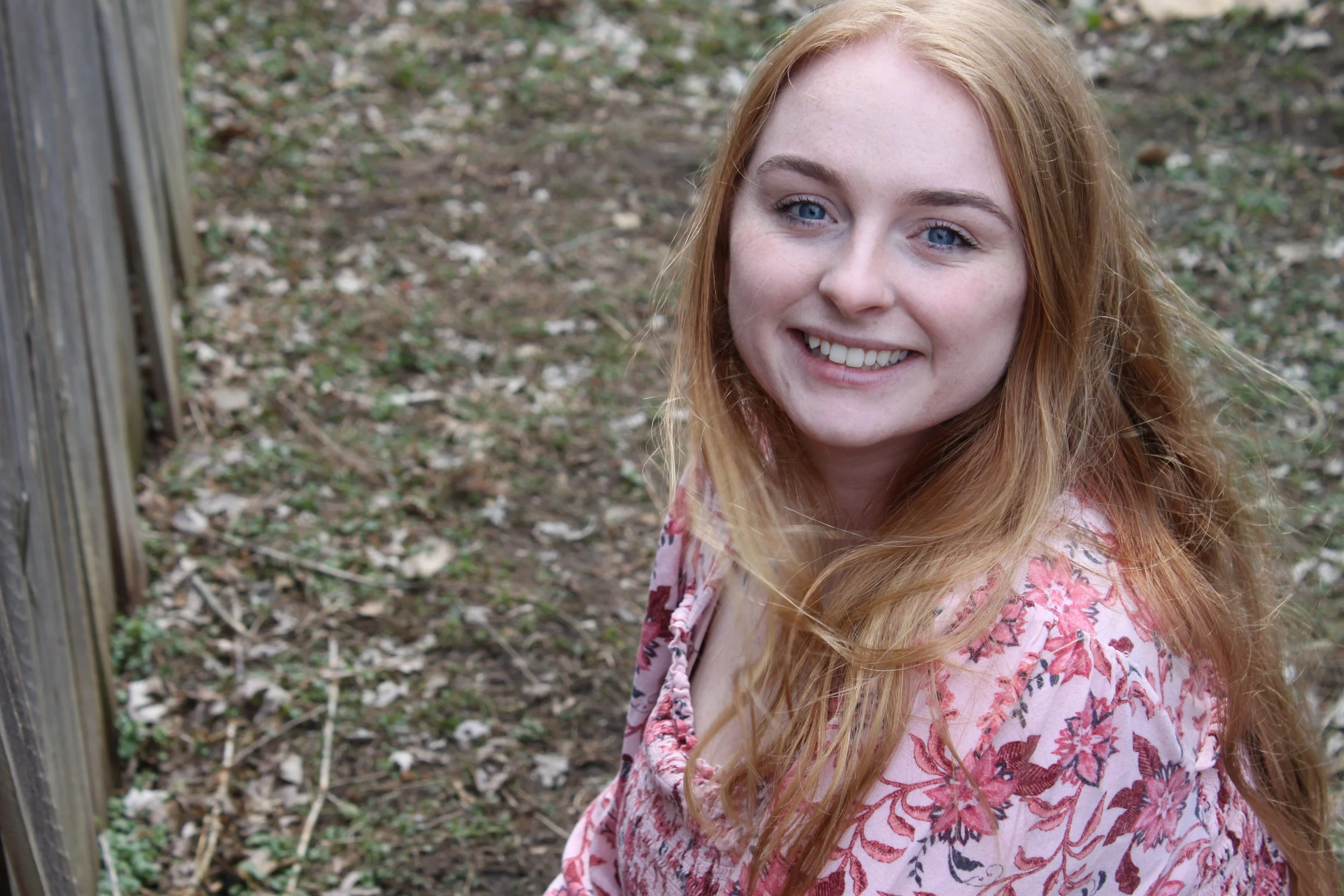A young woman with red hair, blue eyes, and freckles smiling outdoors next to a wooden post, with fallen leaves and grass in the background.