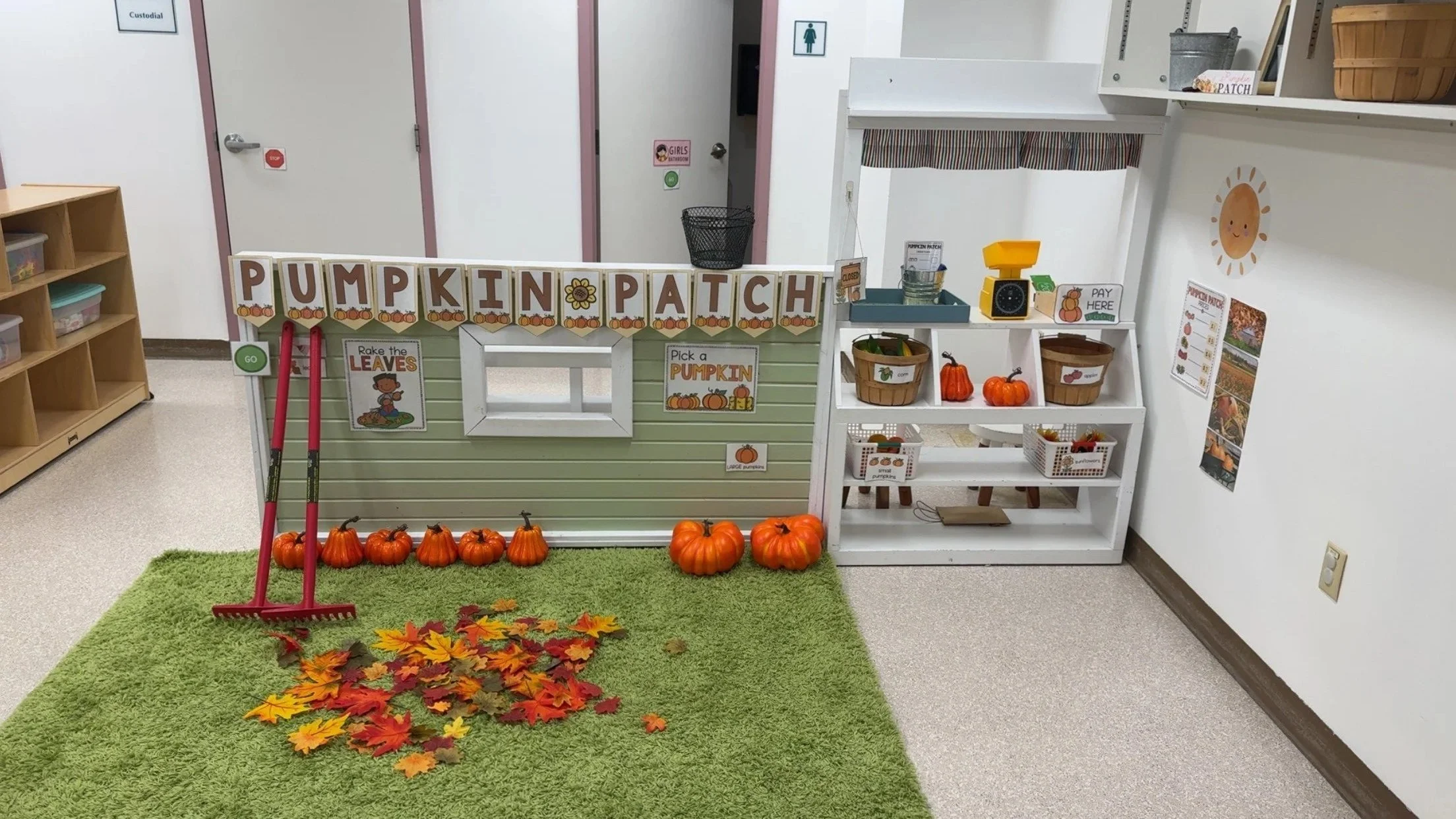 Children's pumpkin patch area with a green rug, fallen leaves, pumpkins, and a small play stand decorated with fall themed signs and baskets of pumpkins.