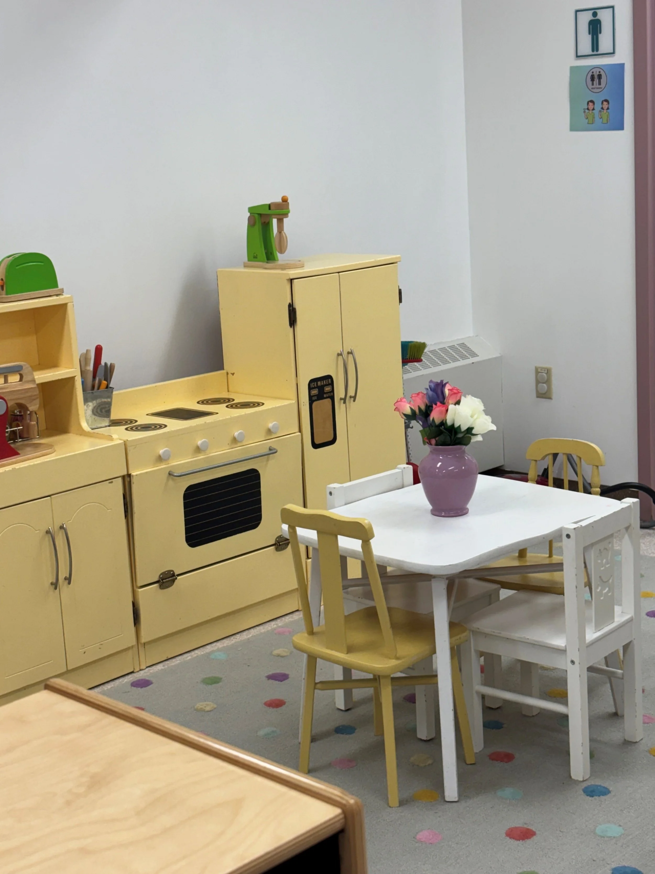 A children's playroom with a small white table and yellow chairs, a yellow play kitchen, and a pink vase with flowers on the table. There are signs on the wall indicating gender and sign language.