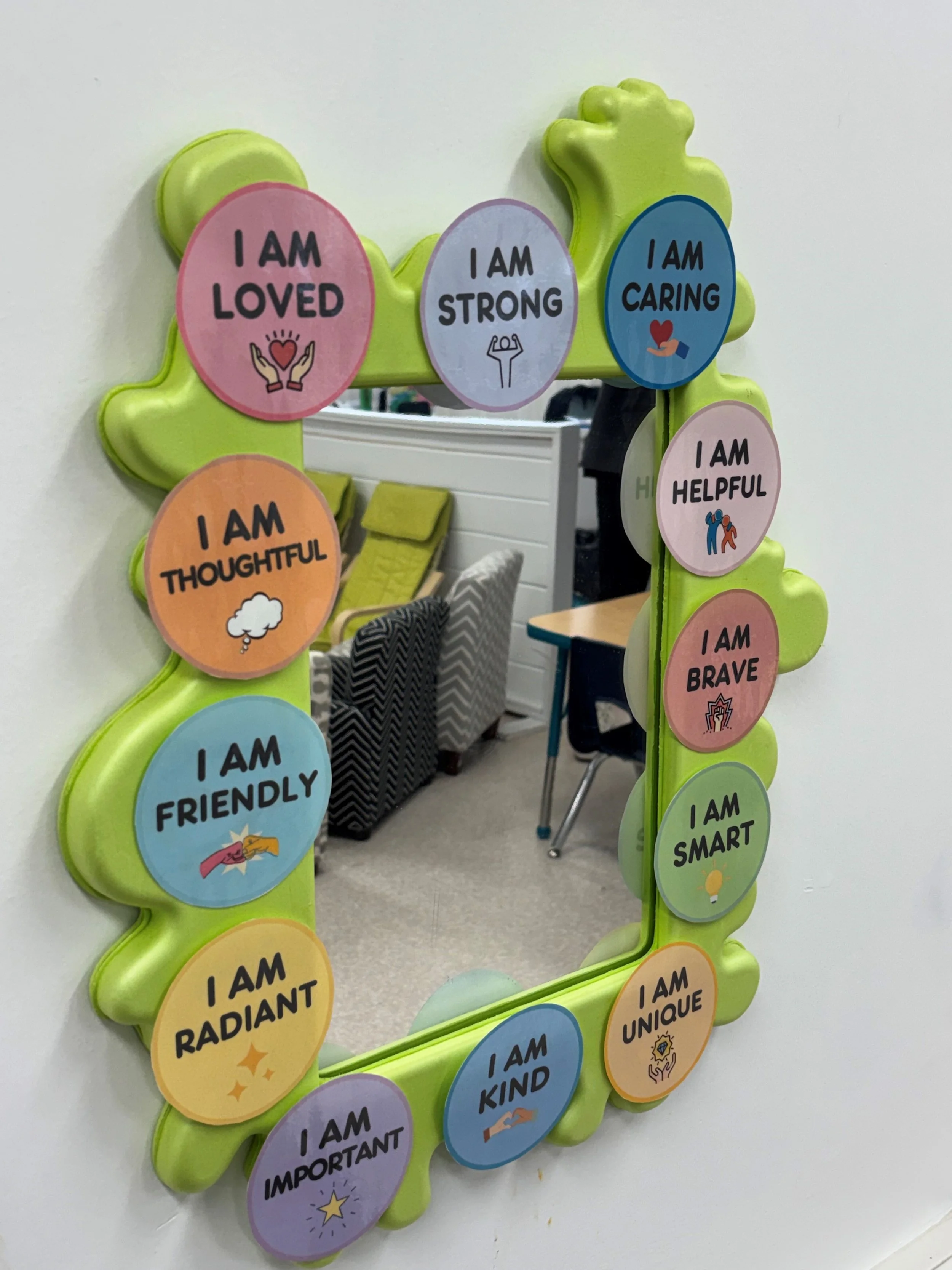 Decorative mirror with circular badges around its border, each with positive affirmations like "I am loved" and "I am brave," on a wall in an office or classroom setting.