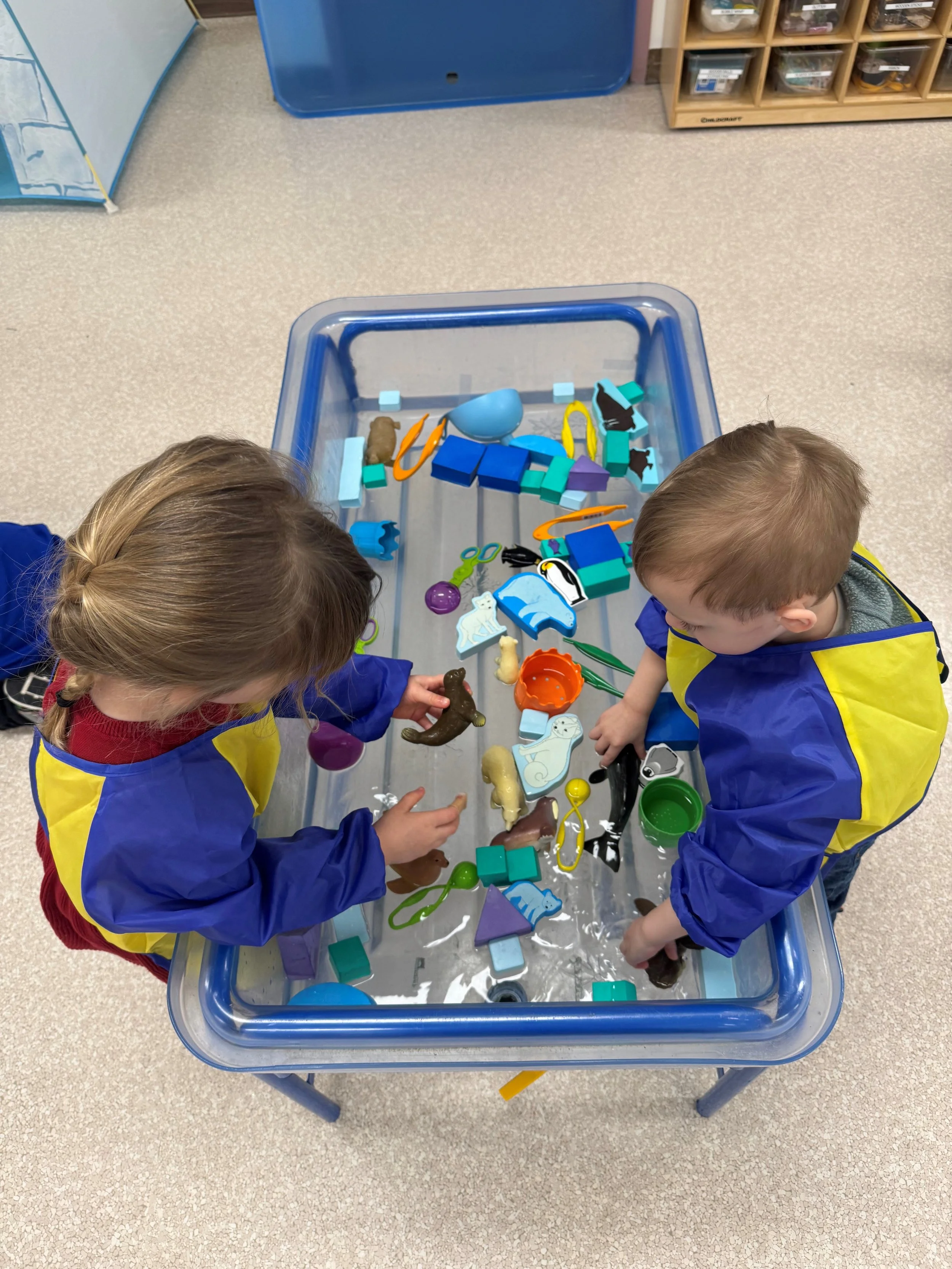 Two children in blue and yellow smocks playing with plastic animal toys in a water-filled sensory bin in a classroom or play area.