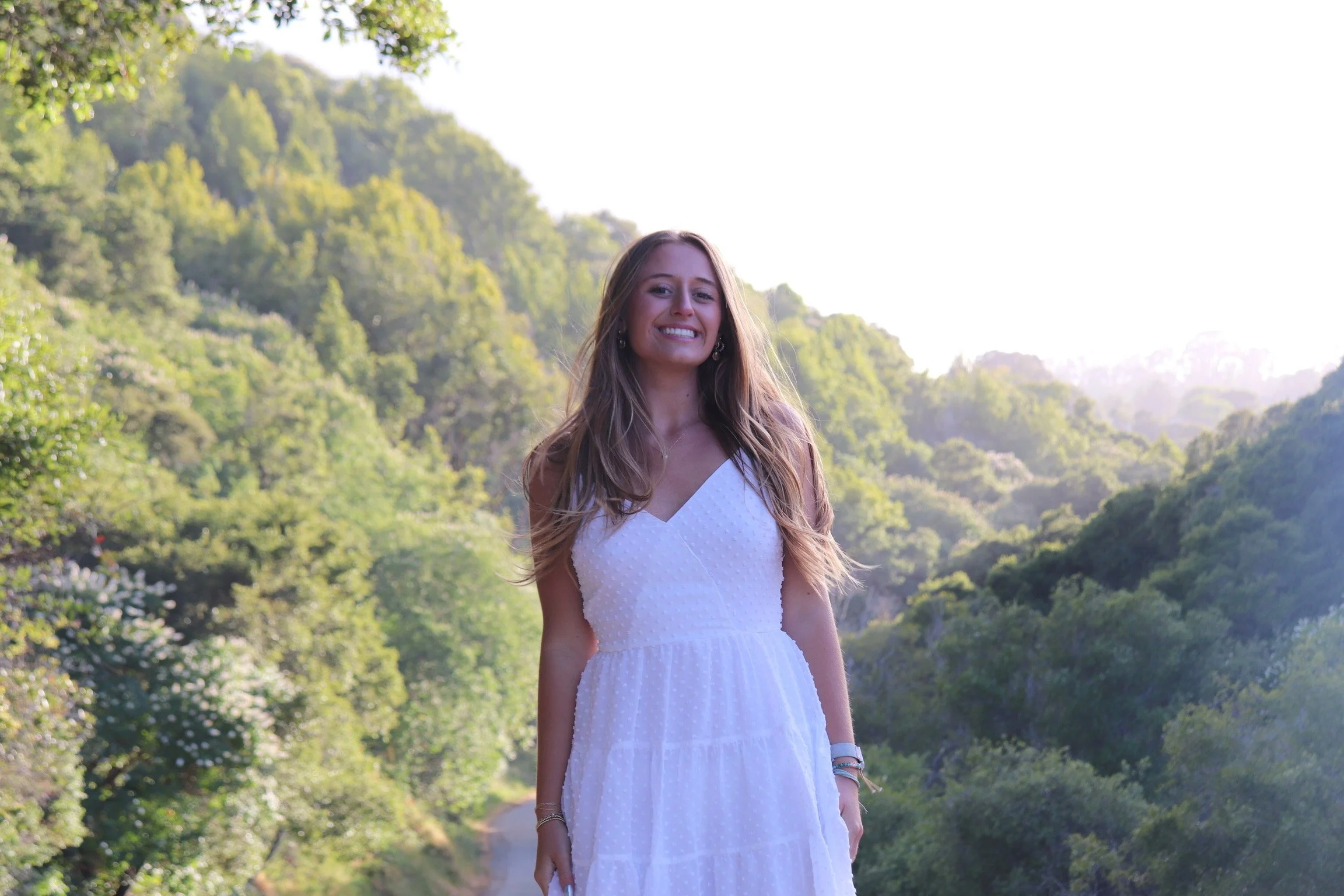 A young woman with long, wavy blonde hair wearing a white sleeveless dress, smiling outdoors with green trees and hills in the background.