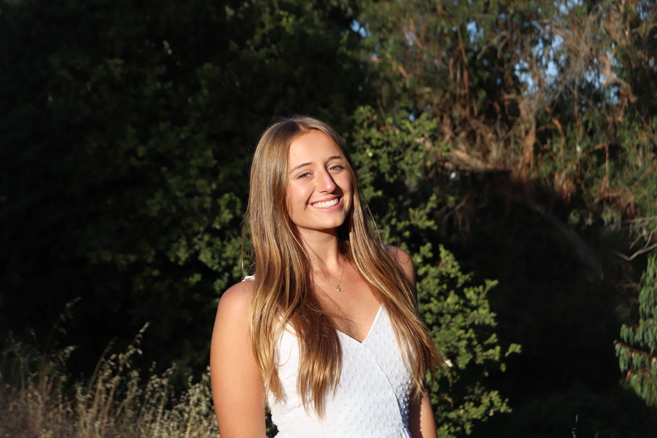 A young woman with long, wavy blonde hair smiling outdoors in sunlight, wearing a white sleeveless top, standing in front of green trees.