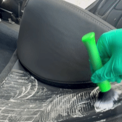 Someone wearing green gloves polishing the back of a car seat with a green applicator pad.