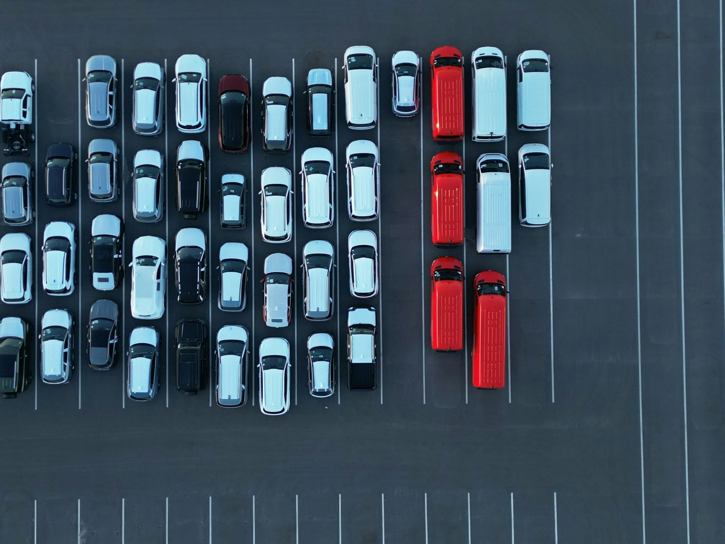 An aerial view of a parking lot filled with white, black, and silver cars and two red buses parked side by side.