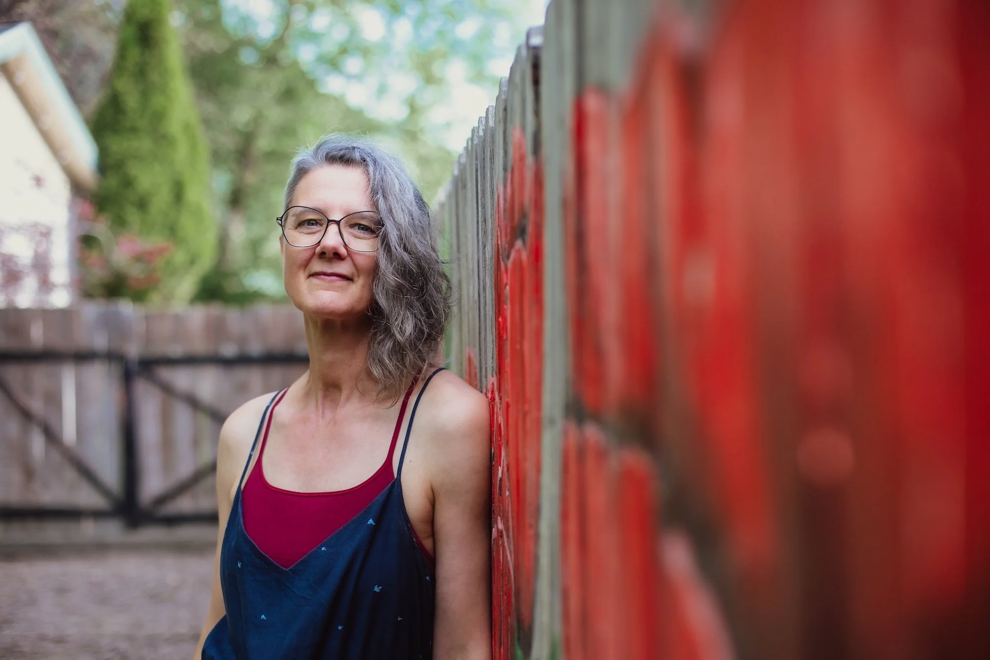 Portrait of Sylke with gray wavy hair, glasses, wearing a sleeveless top, standing outdoors next to a red fence with trees in the background.