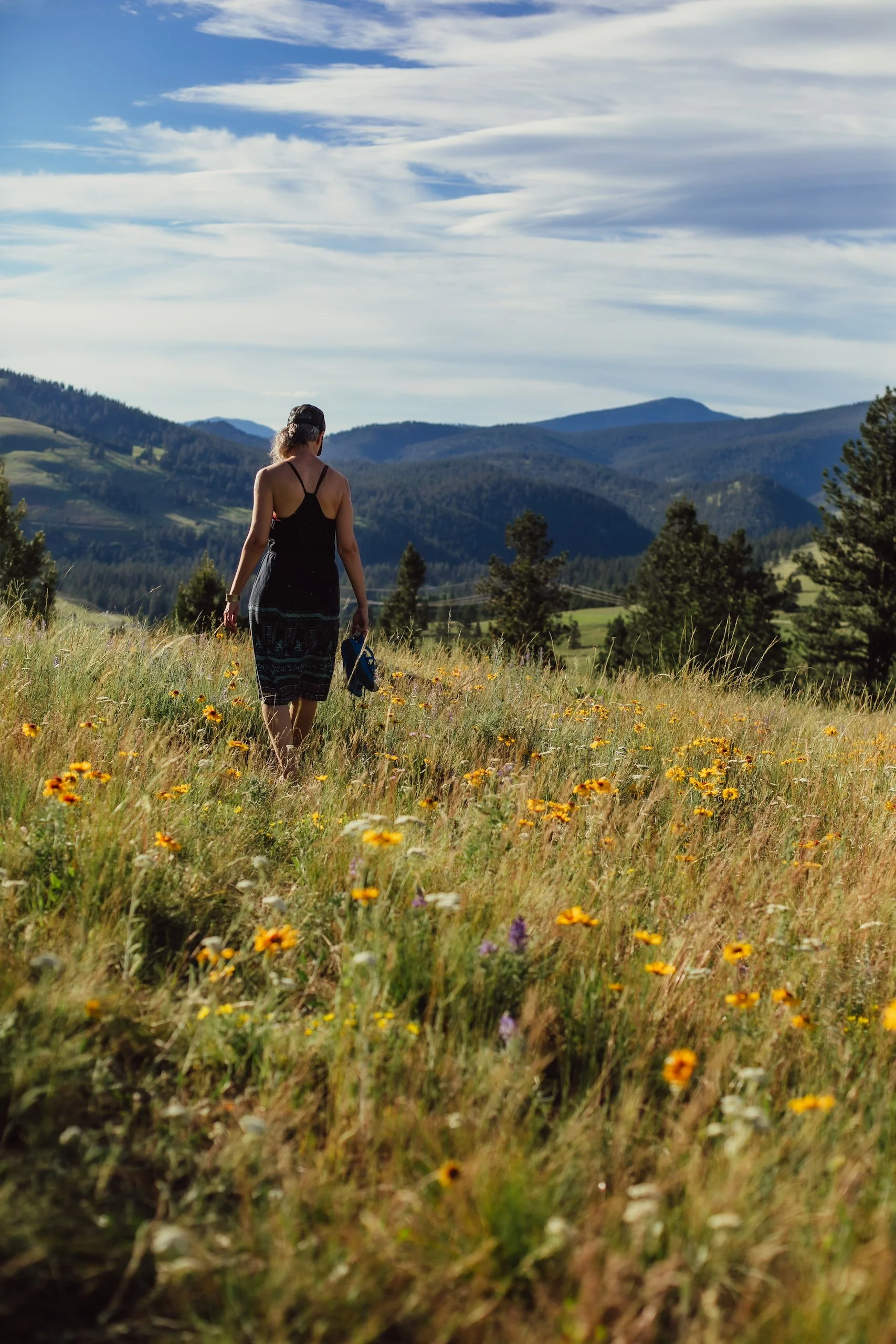A woman walking through a field of yellow and purple wildflowers with mountains in the background under a partly cloudy sky.