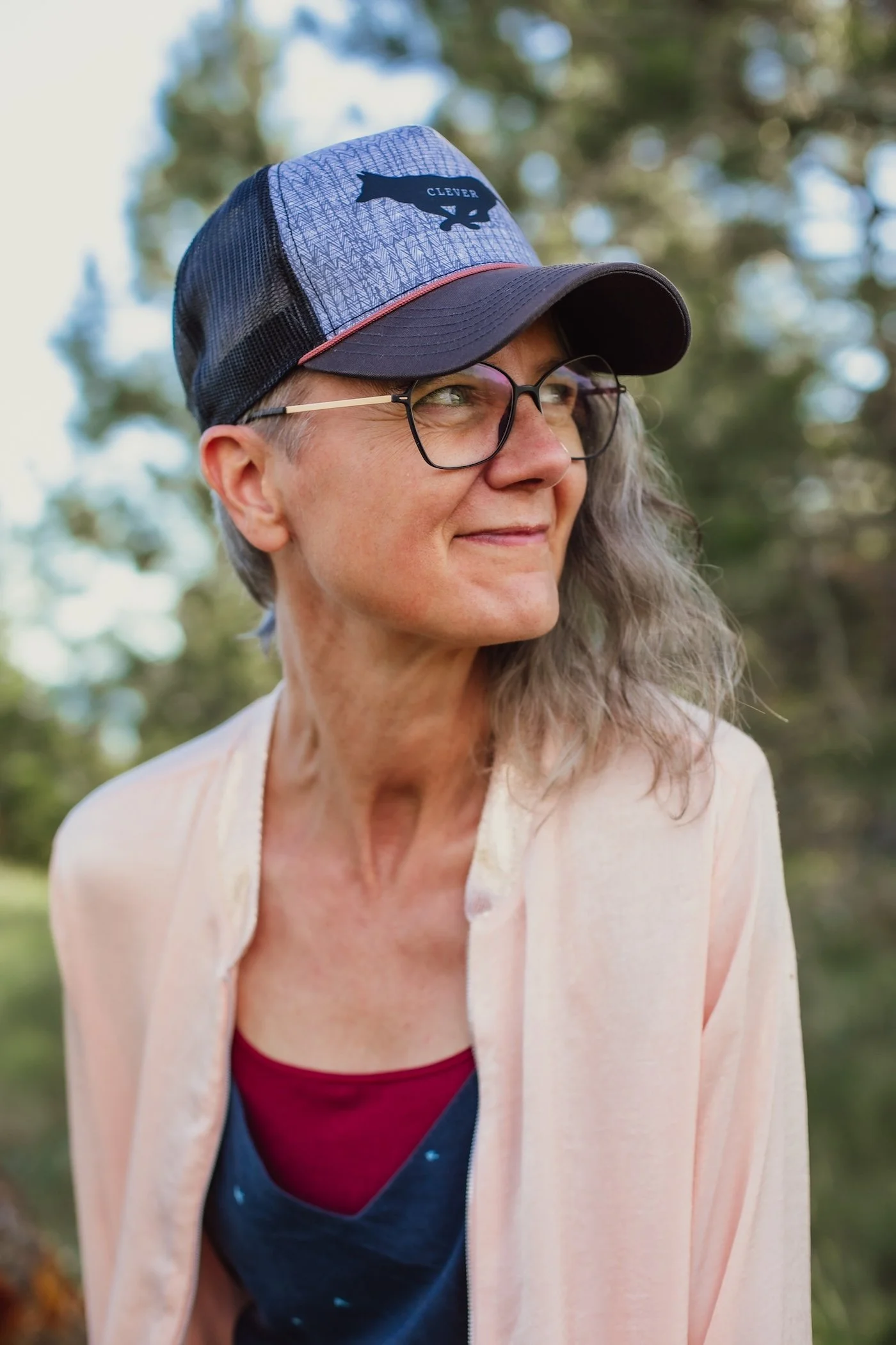 A woman with glasses and long gray hair wearing a blue and gray trucker hat with a black patch that says Clever, a pink cardigan, and a navy top. She is outdoors with blurred trees in the background.
