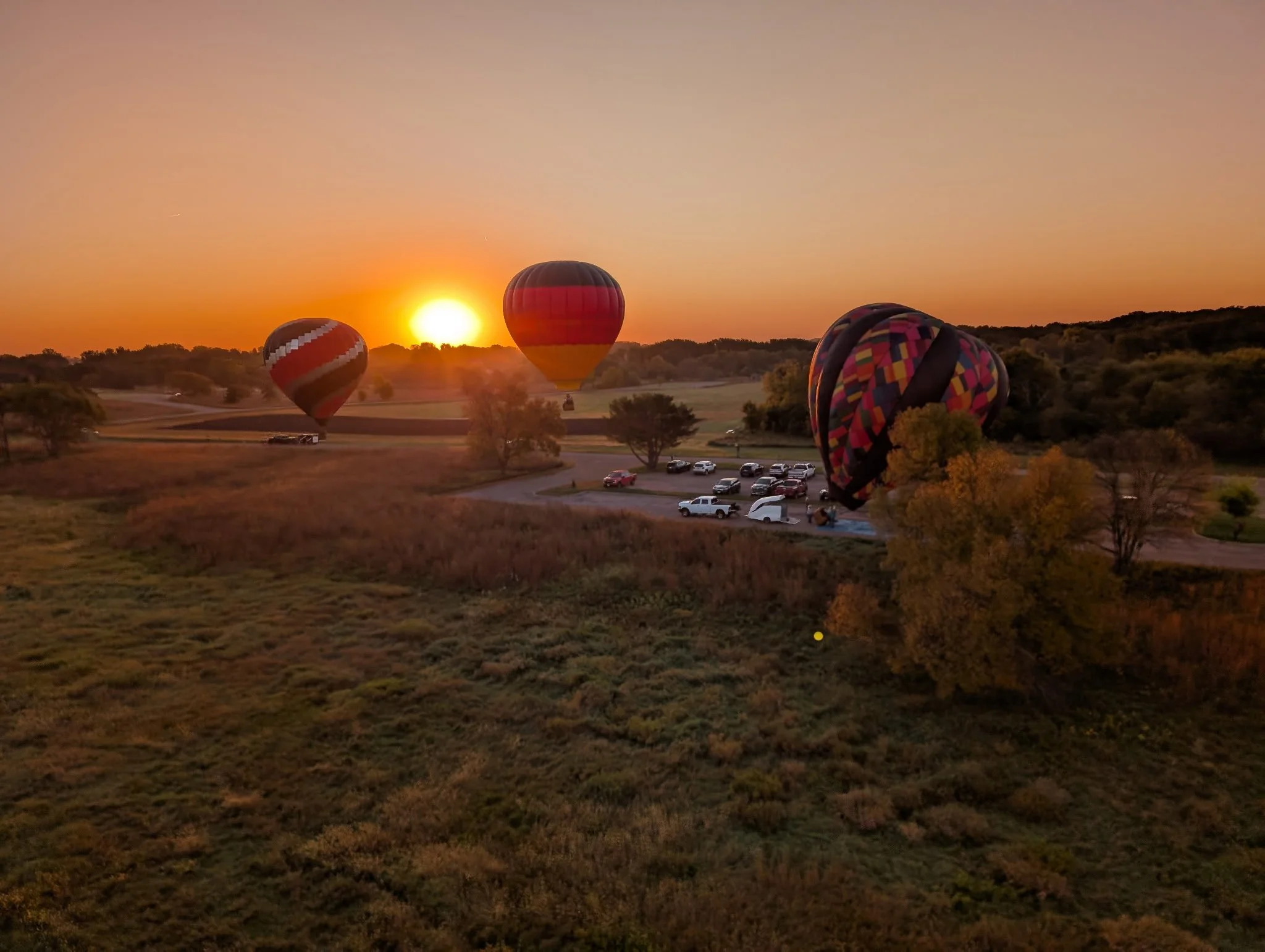 Three hot air balloons floating over a grassy field at sunrise, with parked cars and trees in the background.