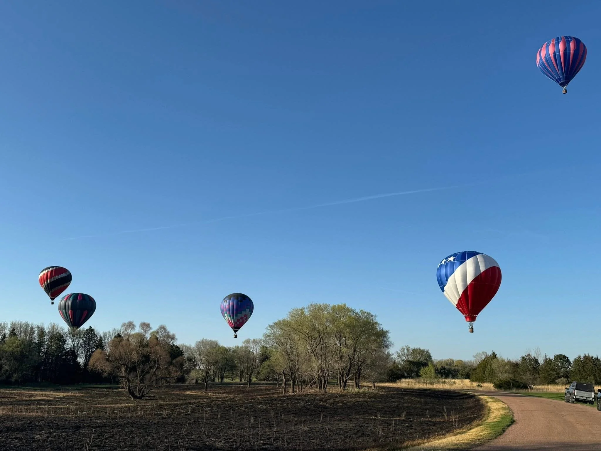 Five hot air balloons floating in a blue sky over a rural landscape with trees and a dirt road.