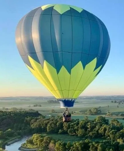A hot air balloon flying over a landscape with trees and fields during sunset or sunrise.