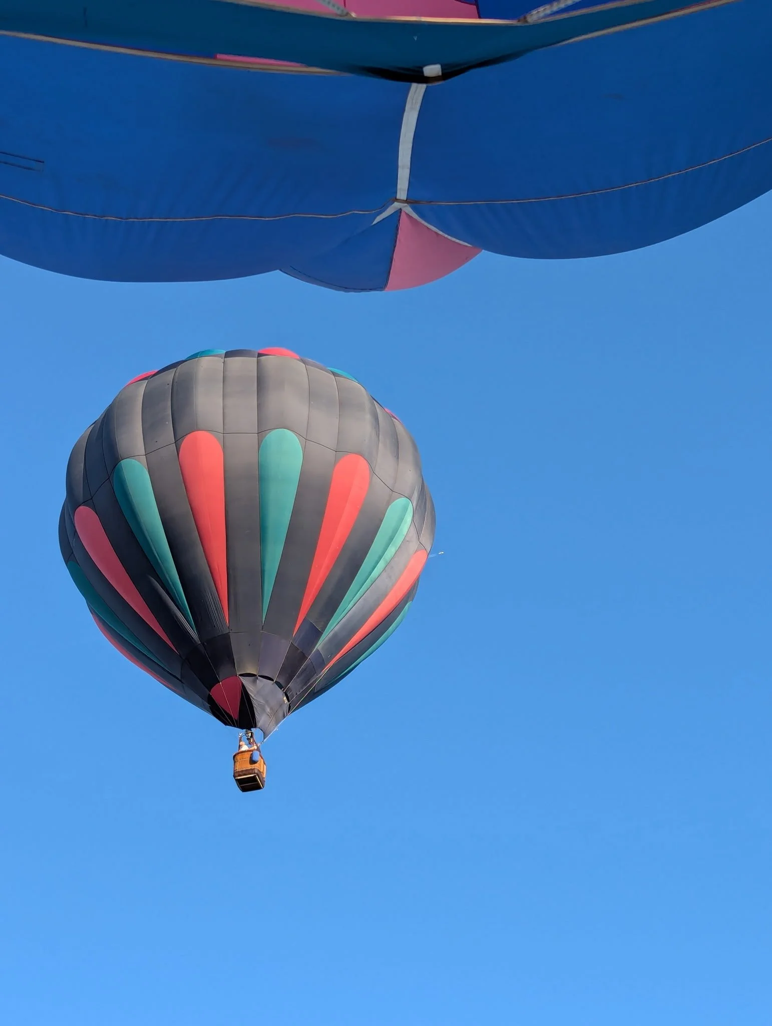 A hot air balloon with black, red, and teal colors floating in a clear blue sky, with the top portion of another balloon visible at the top of the image.