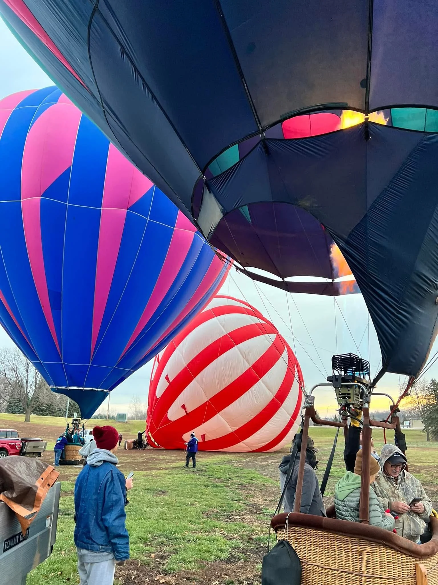Hot air balloons being prepared for takeoff at sunrise, with people on the ground managing the balloons and one person looking at their phone.
