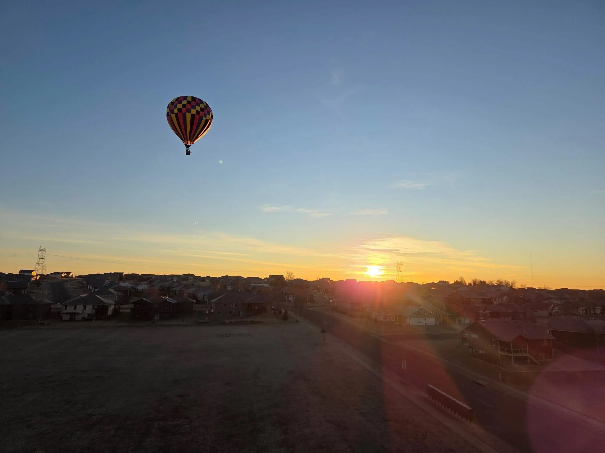 A hot air balloon with a checkered pattern in black, red, and yellow floats in a clear sky during sunset above a suburban neighborhood.