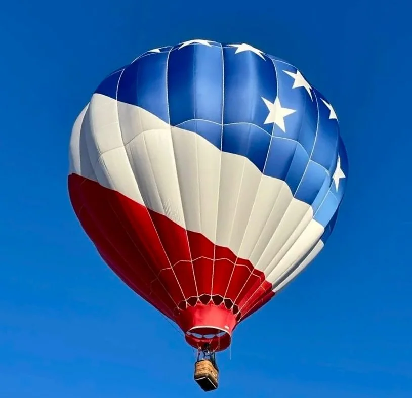 Hot air balloon with American flag design, featuring blue with white stars and red and white stripes, floating in a clear blue sky.