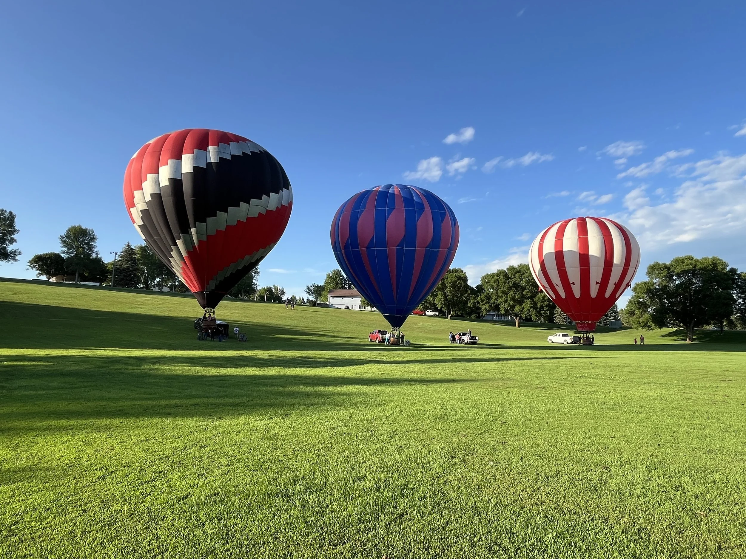 Three hot air balloons in the sky above a grassy field, with trees and a few people visible in the background on a clear day with some clouds.