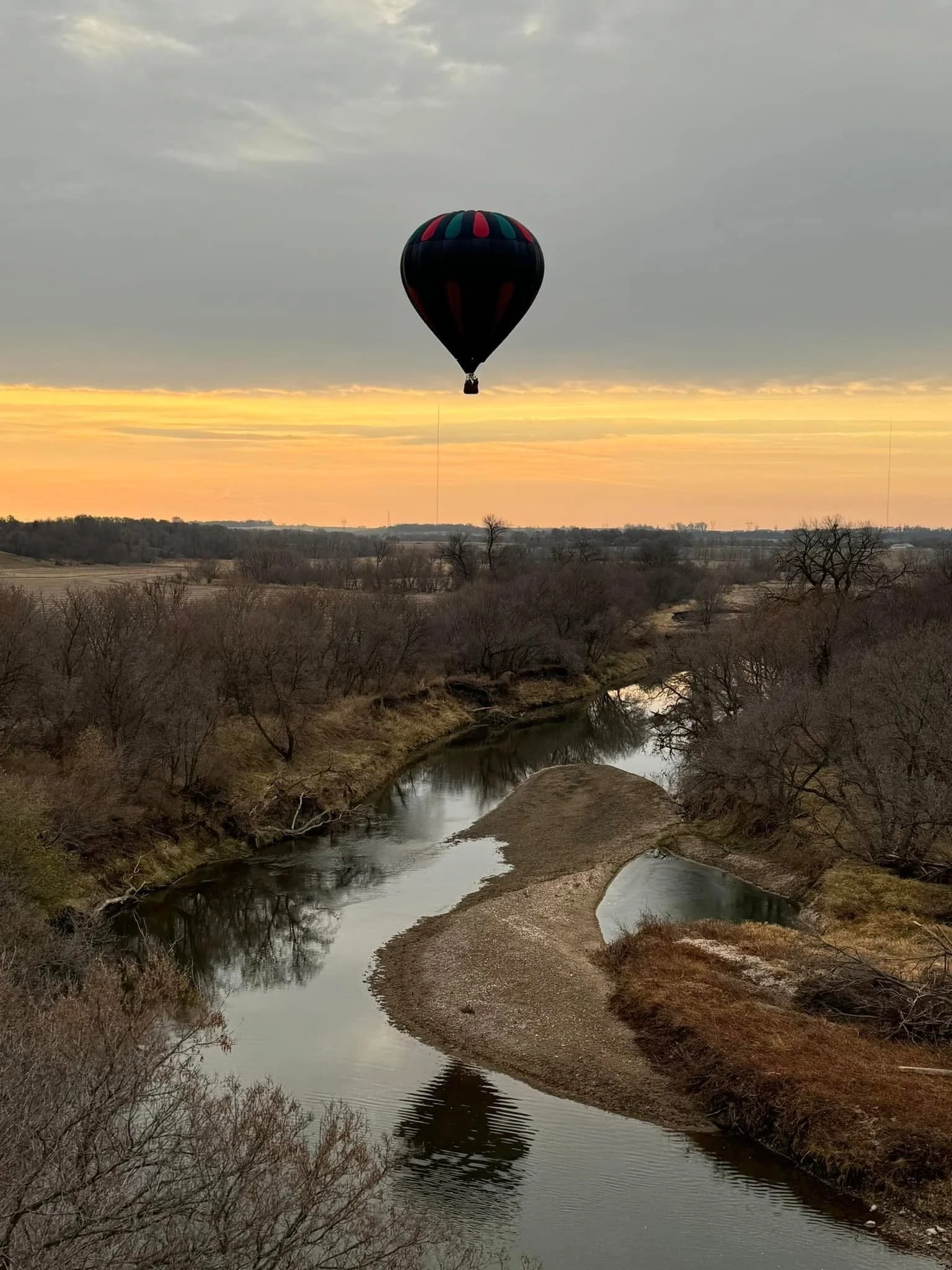 A hot air balloon floating in the sky during sunset over a river with leafless trees along the banks.