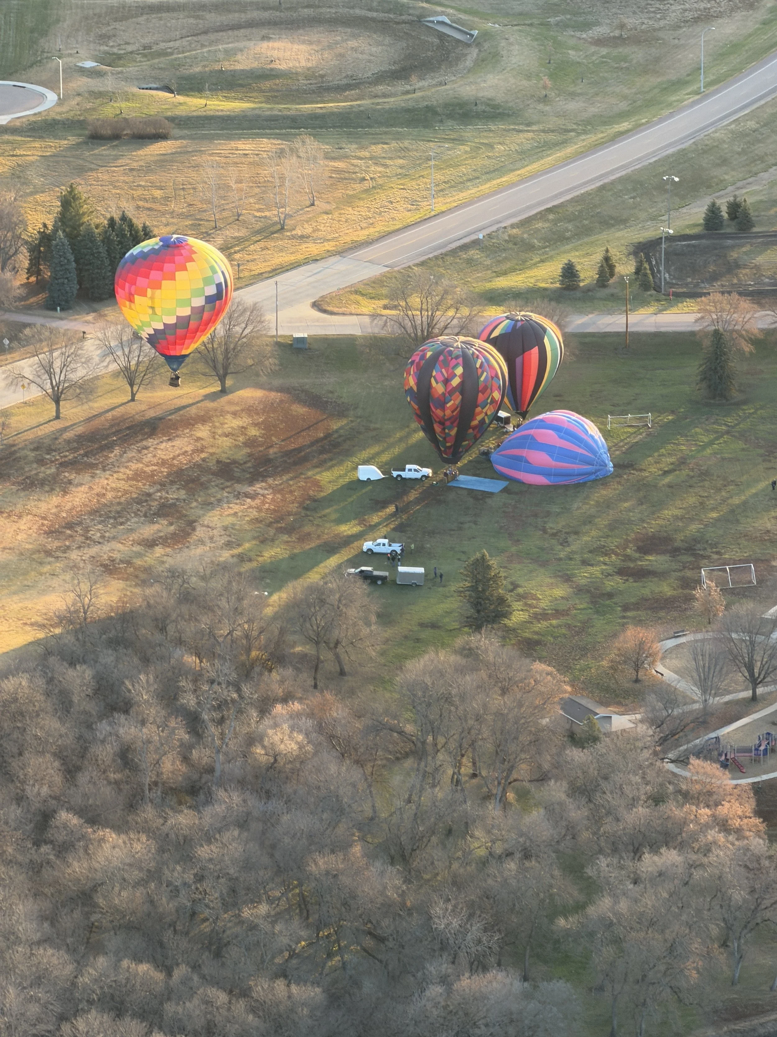 Four colorful hot air balloons floating above a park or open field with trees, playground, and pathways, during sunset or sunrise.