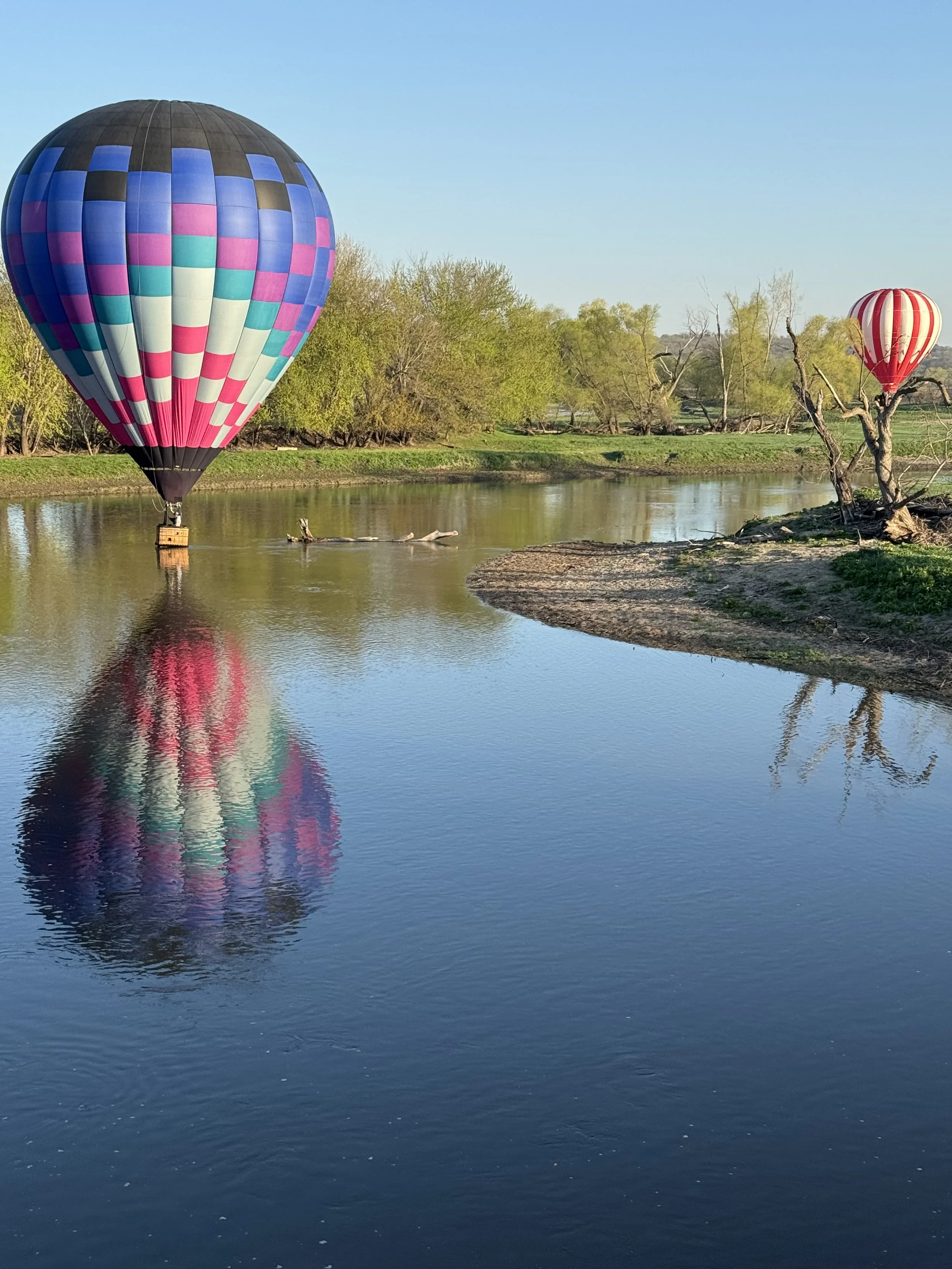 Two colorful hot air balloons floating over a calm river, with lush green trees and a clear blue sky in the background.