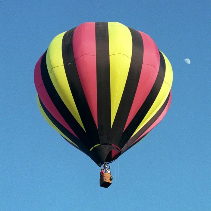 Colorful hot air balloon with pink, yellow, and black stripes flying in a clear blue sky, with the moon visible in the background.