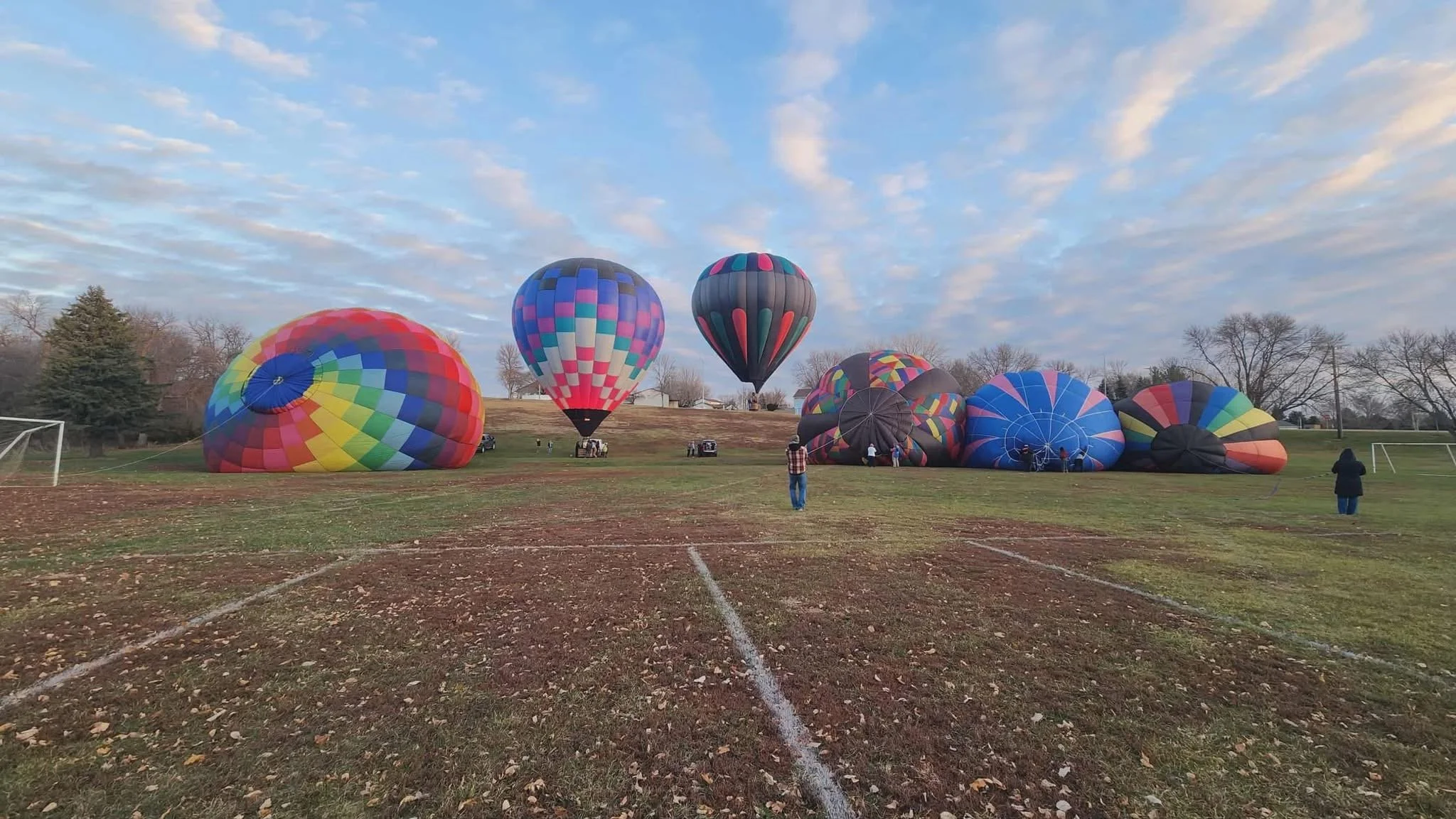 Several colorful hot air balloons on a grassy field, some are partially inflated and others upright, with a few people standing nearby.