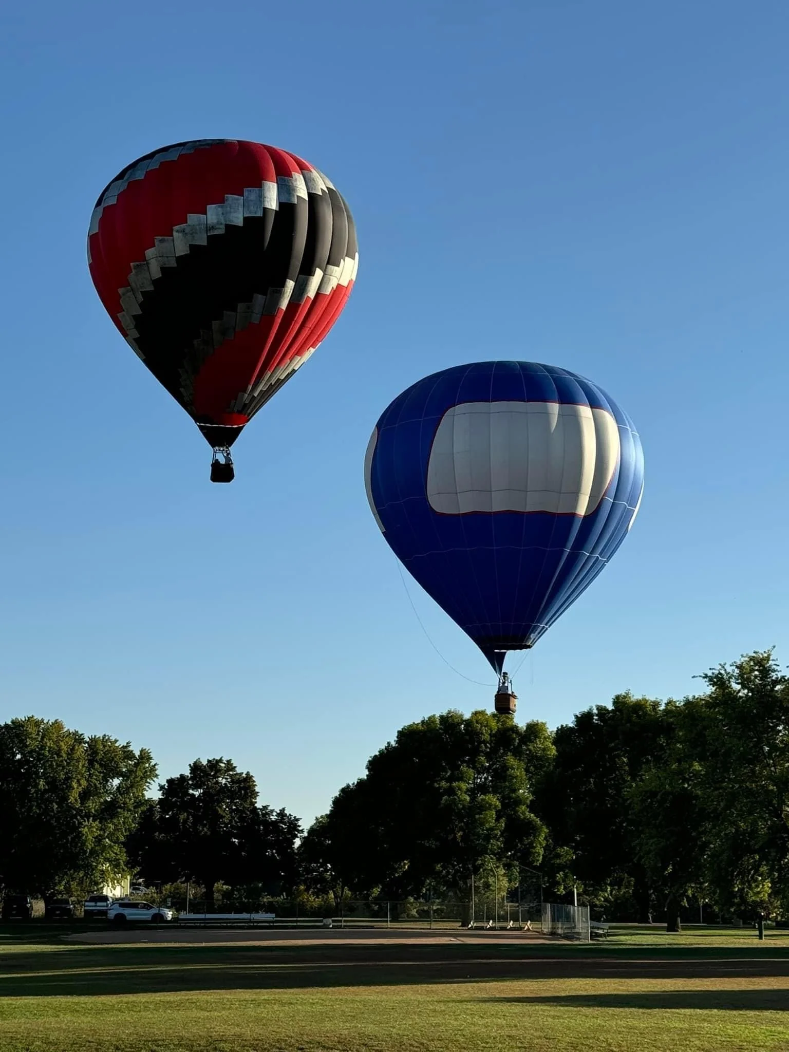 Two hot air balloons floating in clear blue sky above a park with trees and a baseball field.