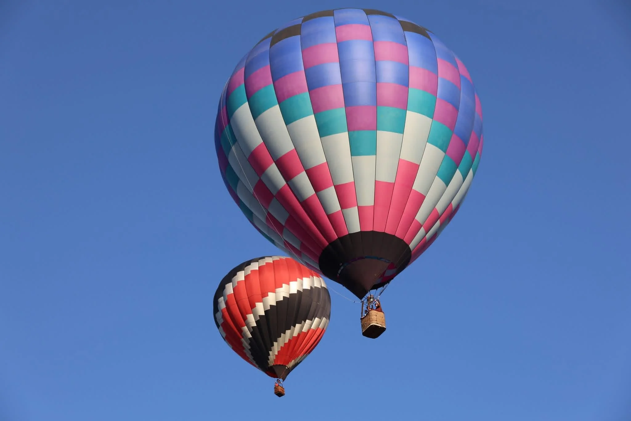 Two colorful hot air balloons floating in a clear blue sky, one larger with pink, white, and blue checkered pattern, and one smaller with red, black, and white pattern.