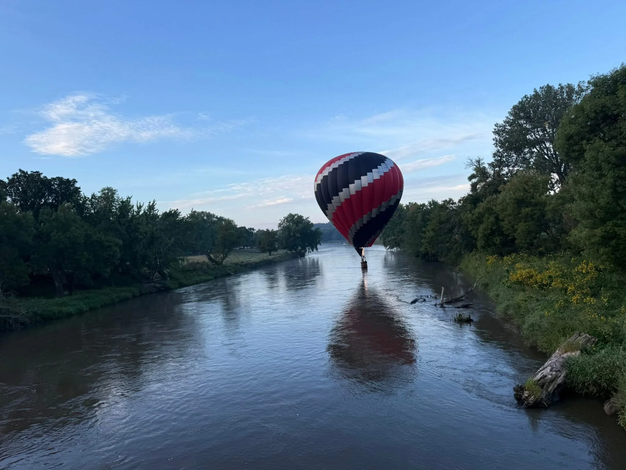 A hot air balloon with black, white, and red stripes floating over a river, with trees lining the riverbanks under a blue sky with some clouds.