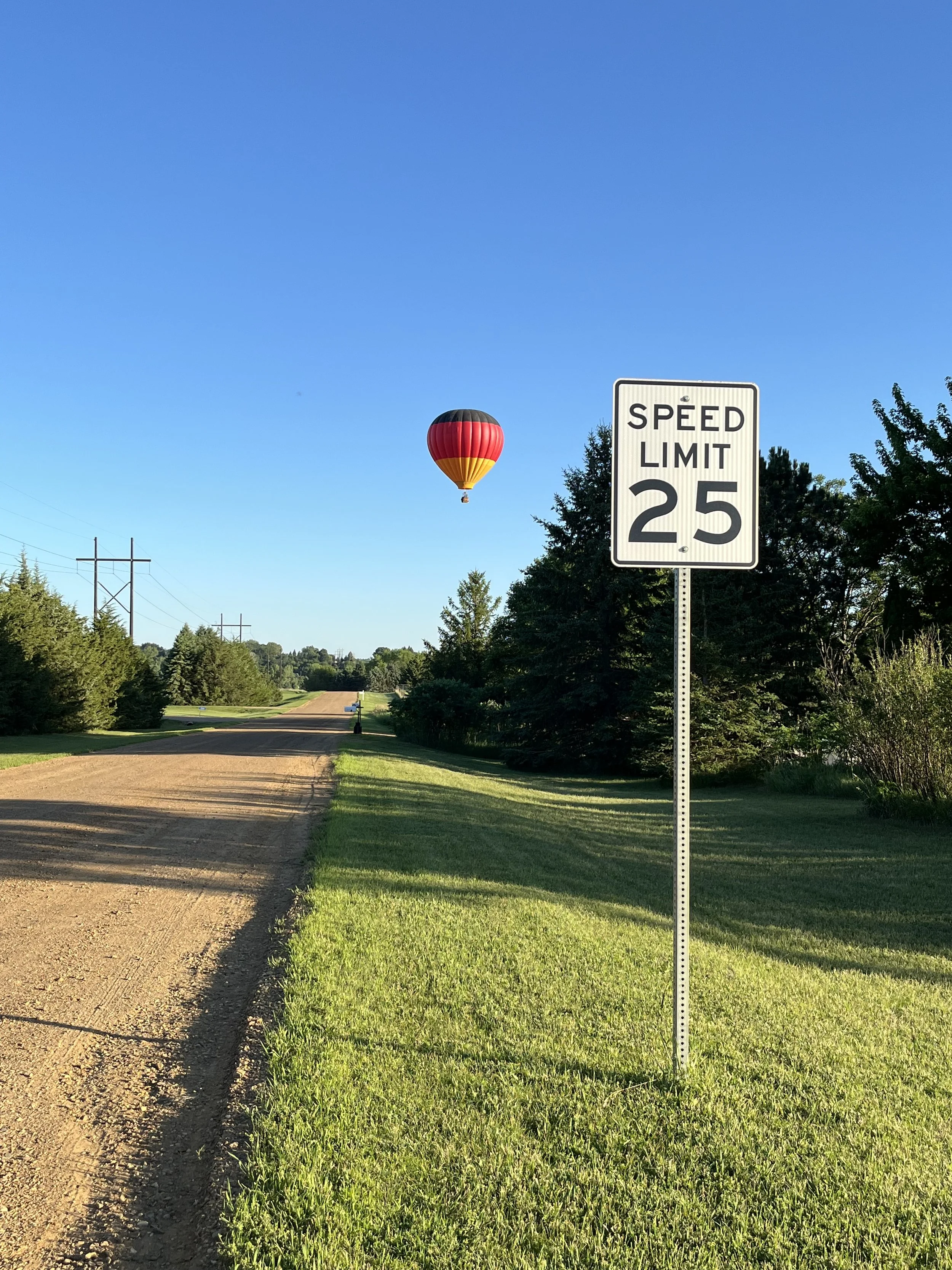 A gravel and dirt road with a speed limit sign of 25 mph on the right side, green grass on both sides, trees lining the road, a hot air balloon floating in a clear blue sky in the background, and power lines running parallel to the road.