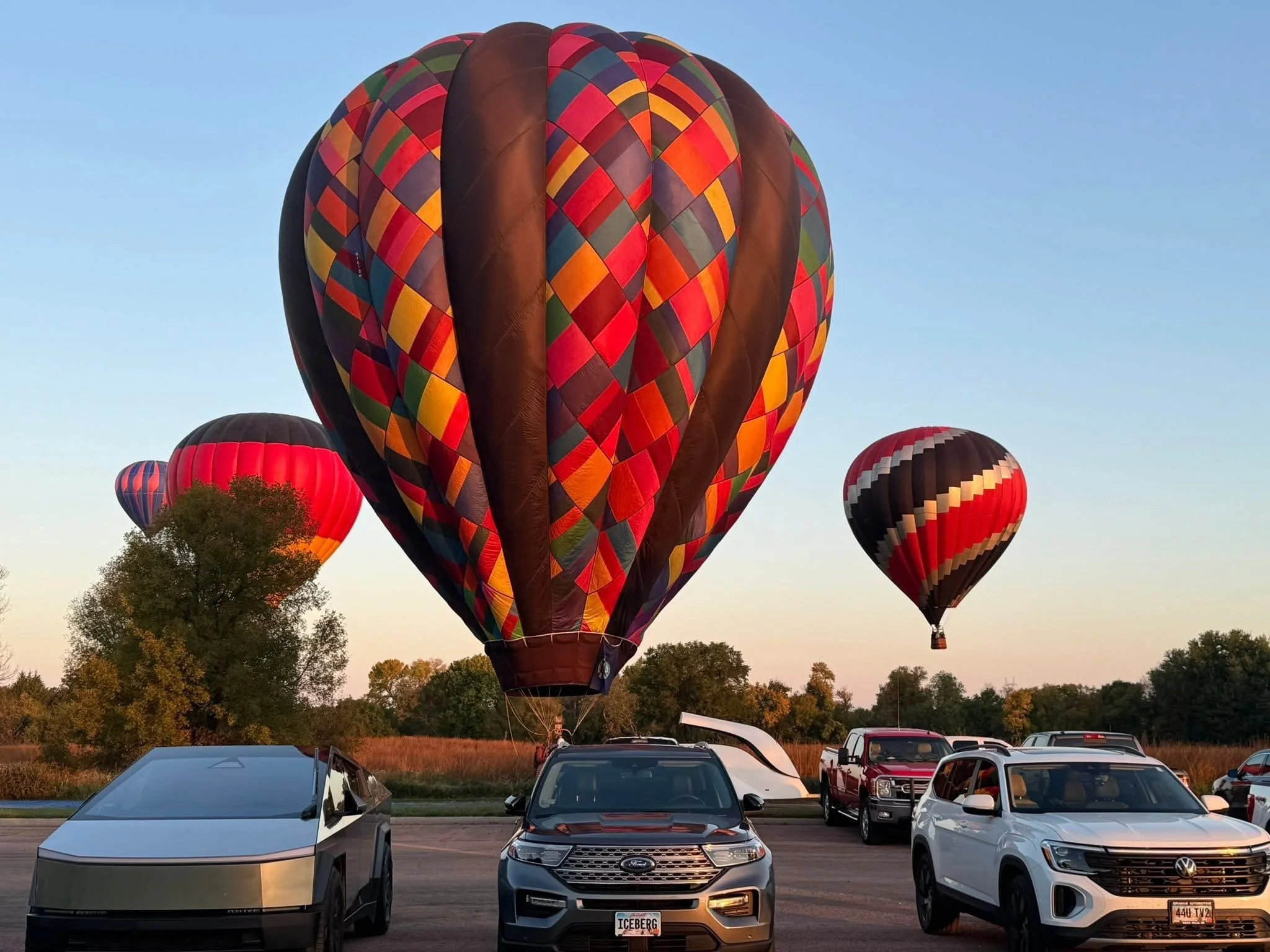 Multiple hot air balloons are floating in the sky near parked cars on the ground during sunset.