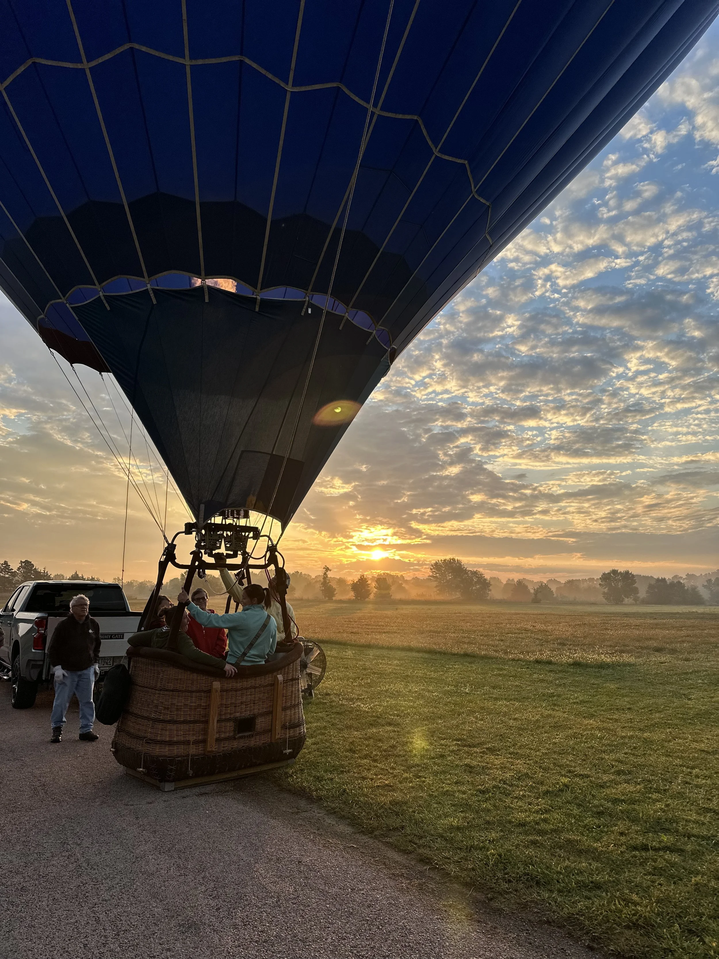 Hot air balloon preparing for takeoff during sunrise with several people inside the basket and a vehicle nearby on a grassy field.