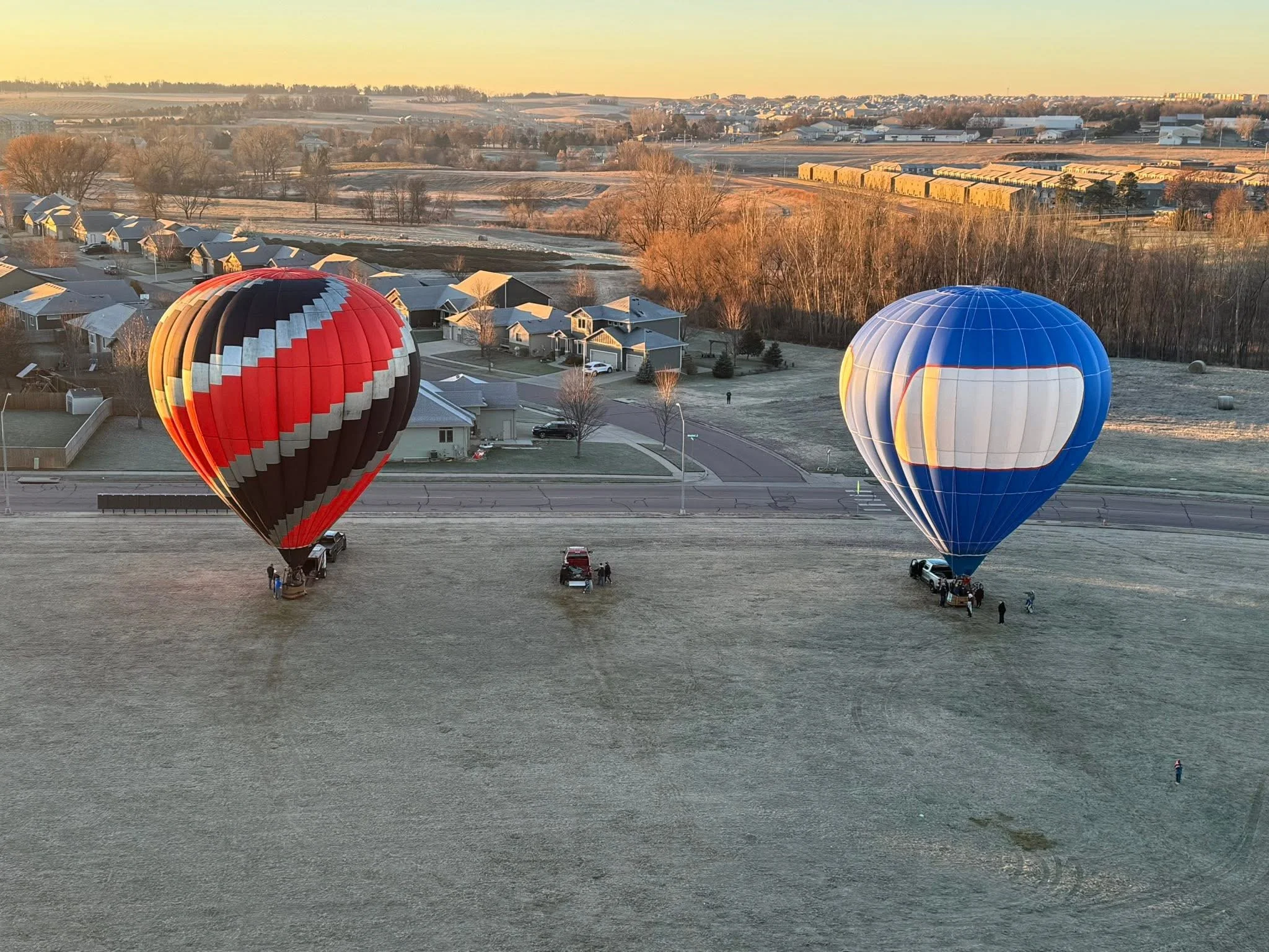 Two hot air balloons, one red and black striped and the other blue and white, are preparing for flight on an open field during dawn or dusk.