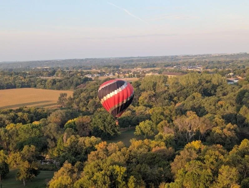 A hot air balloon with red, black, white, and gray stripes floating above a green, tree-filled landscape.