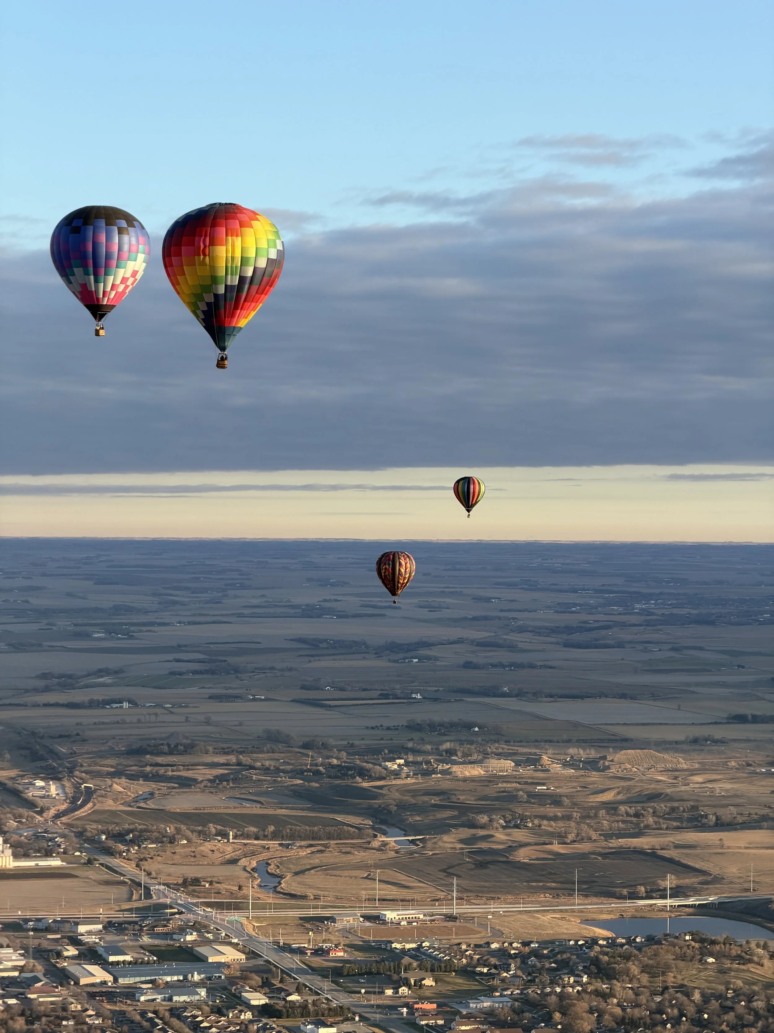 Four colorful hot air balloons floating in the sky over a rural landscape with fields, roads, and buildings.