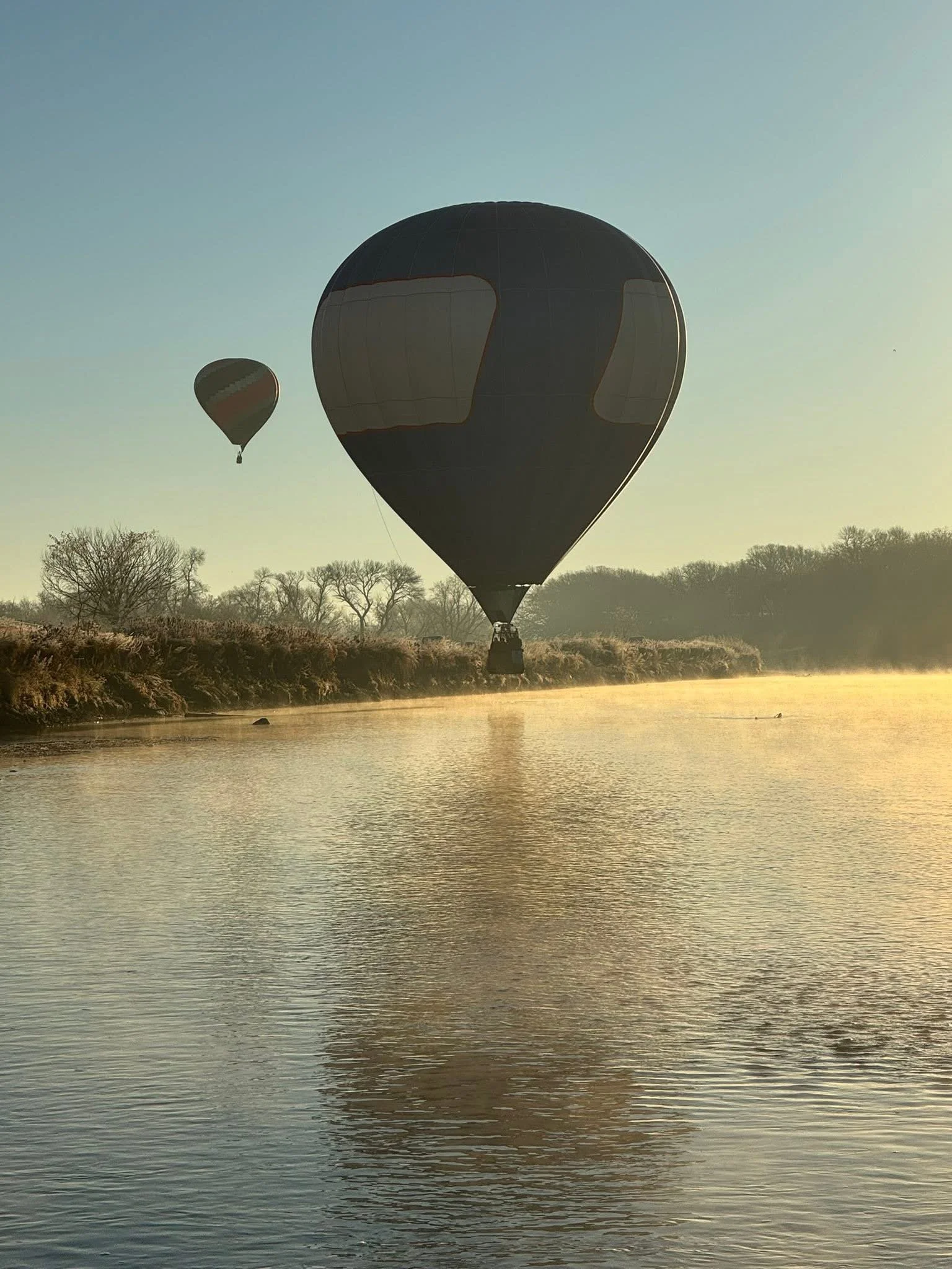 Two hot air balloons floating over a calm river at sunrise, with trees in the background.